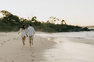 Two people walking together on a sandy beach at sunset with ocean waves and trees in the background.