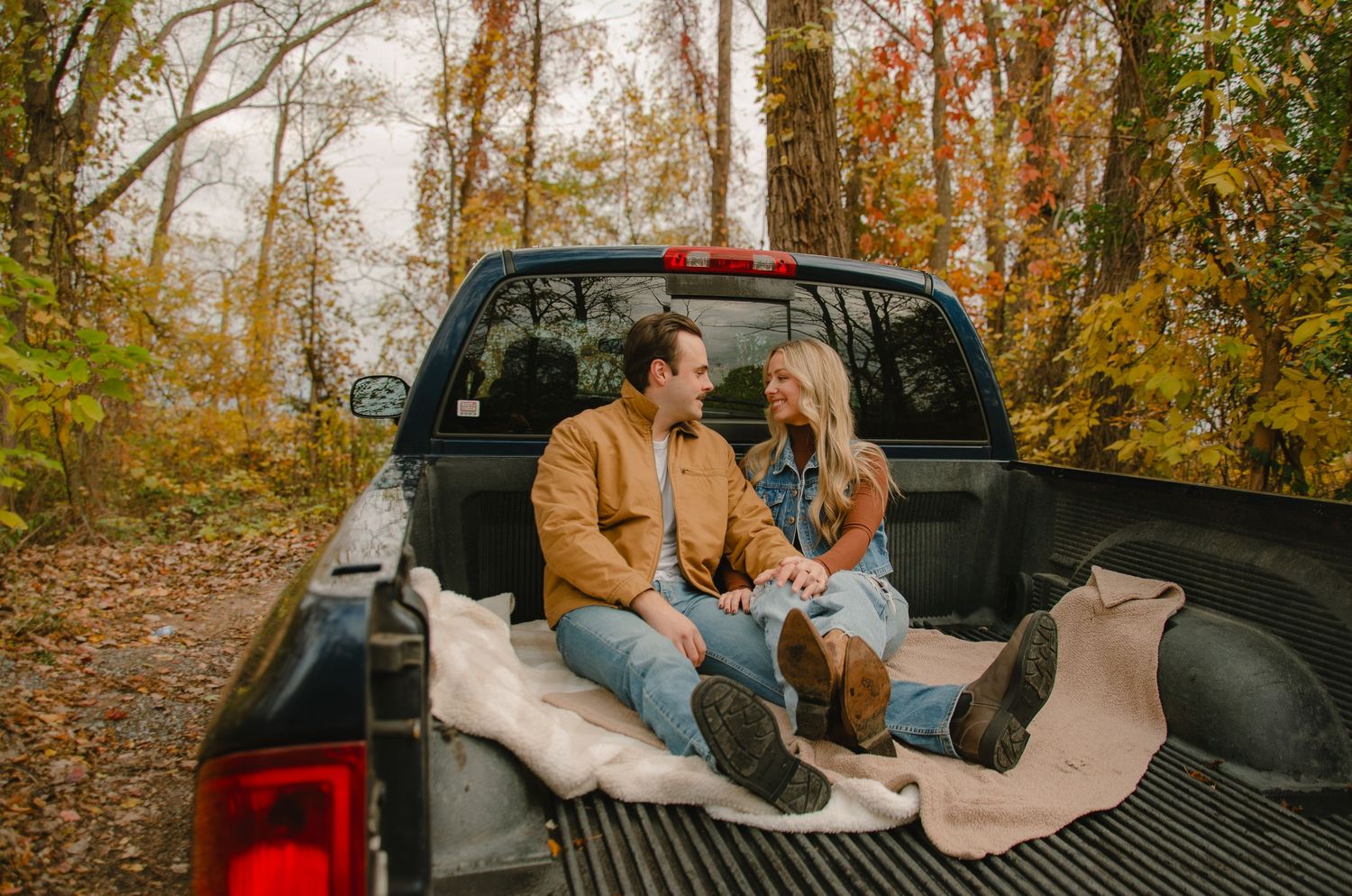 A couple sits in the back of a pickup truck surrounded by autumn foliage in a romantic outdoor setting.