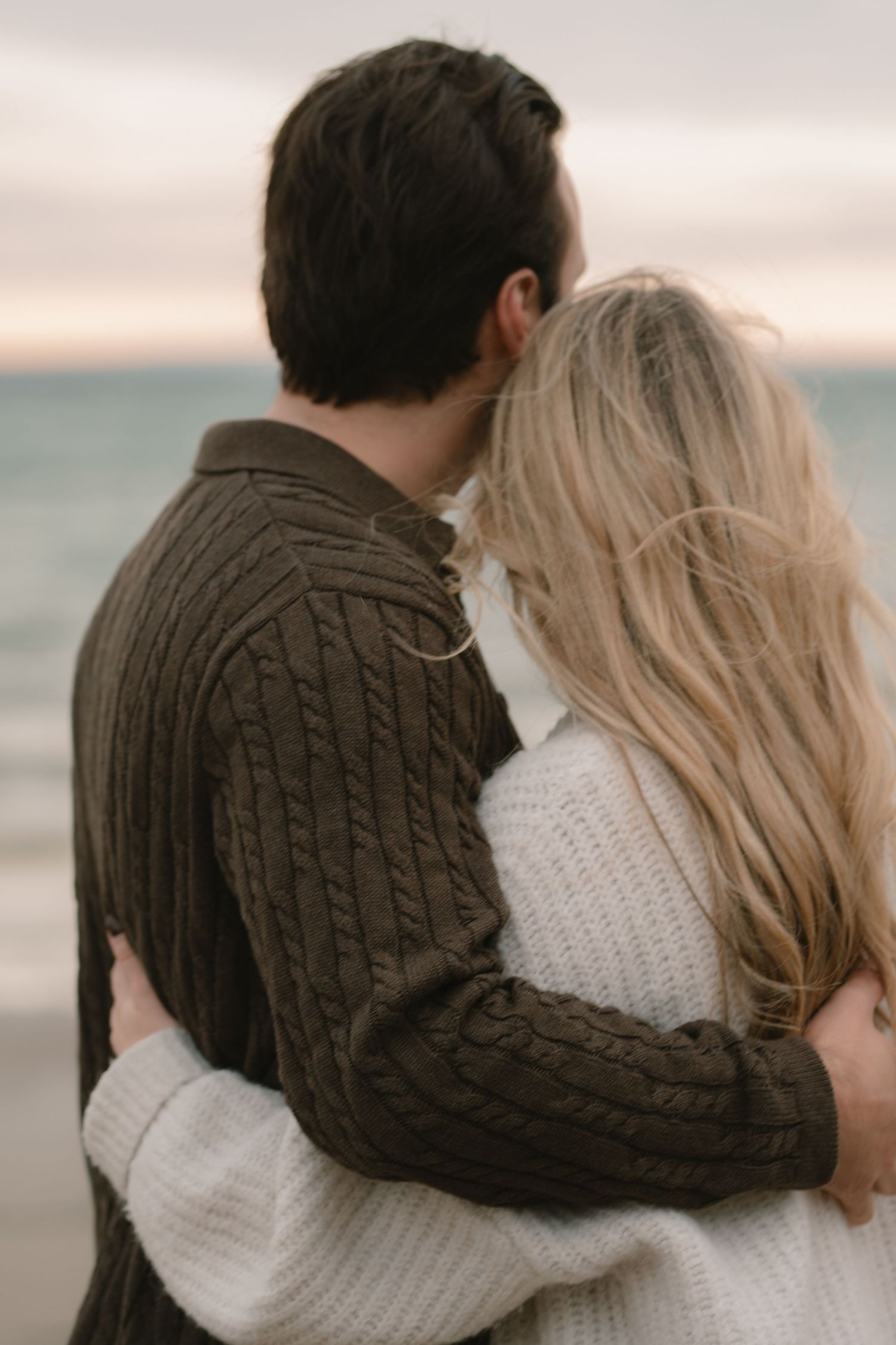 Couple in cozy sweaters embracing while looking at ocean during sunset.