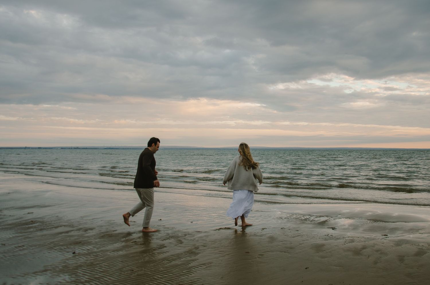Silhouettes walking along a misty beach at sunset with cloudy skies and ocean waves.