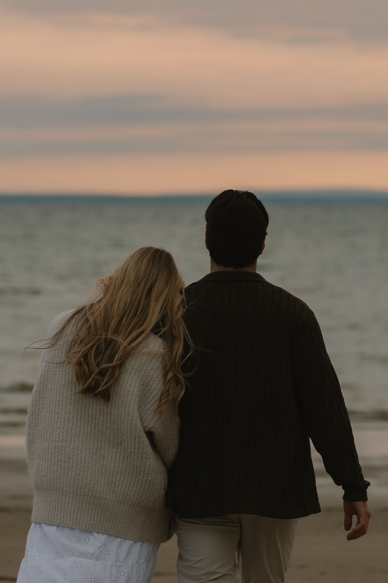 A couple in cozy sweaters walks along the shoreline at sunset.