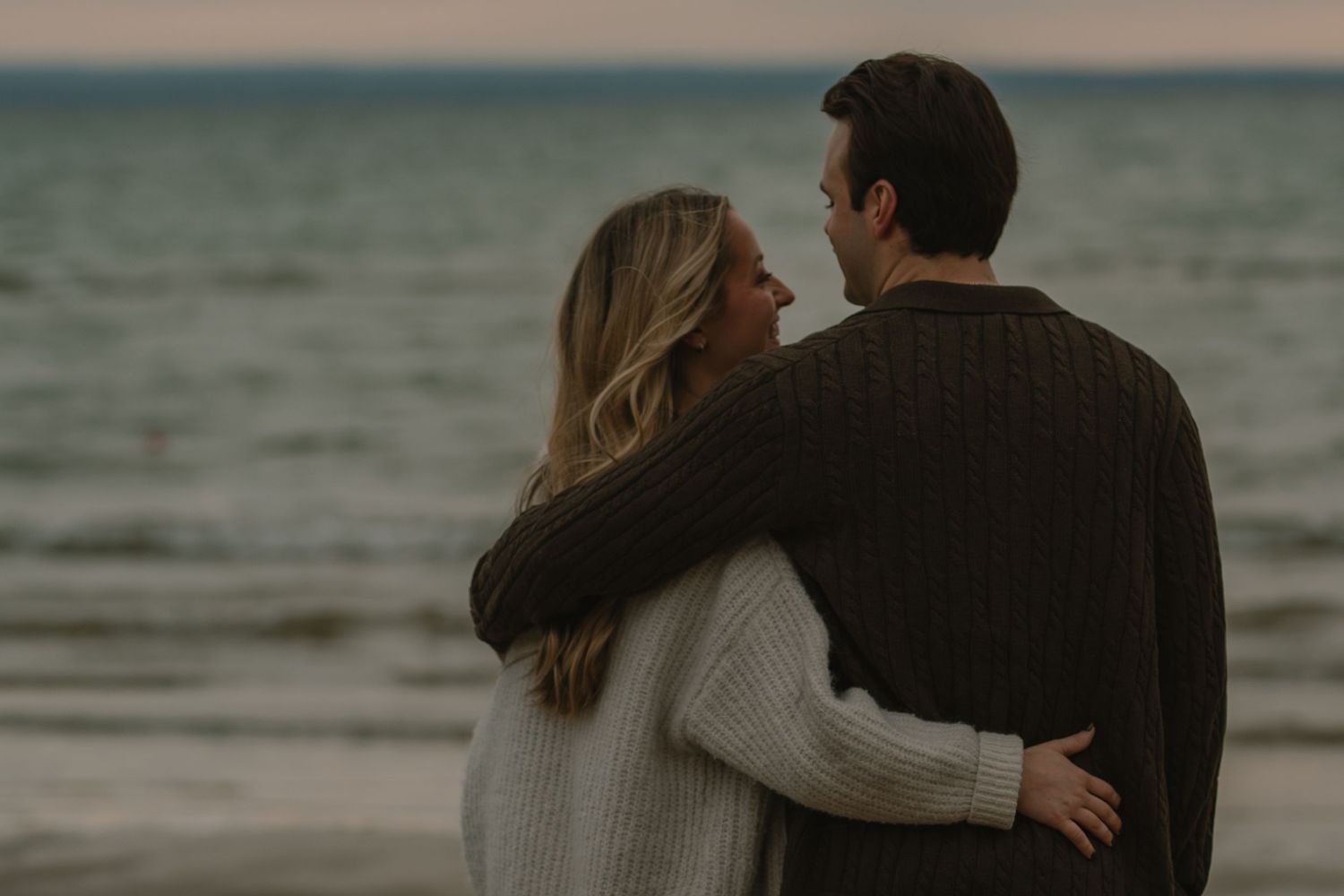 Two people embrace while walking along a misty beach at dusk.