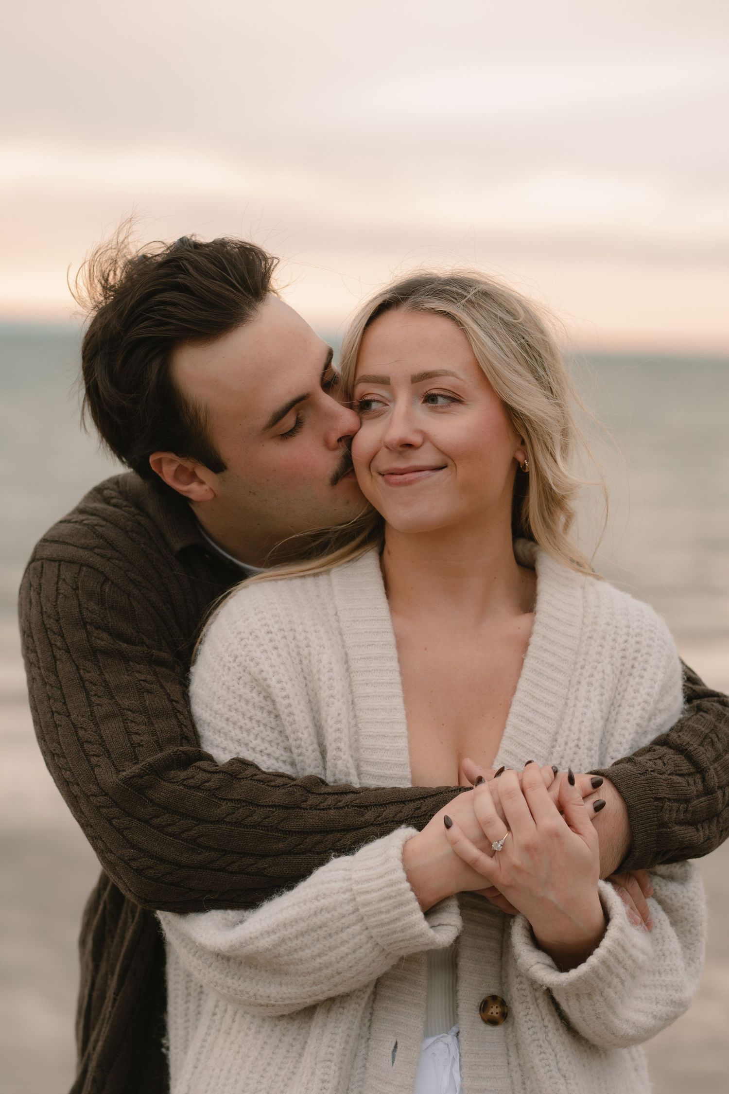 A romantic couple embraces on the beach at sunset while showing off a new engagement ring.
