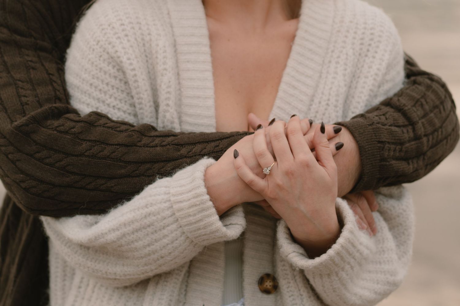 Close-up of couple wearing white and brown knit sweaters holding hands by the sea.