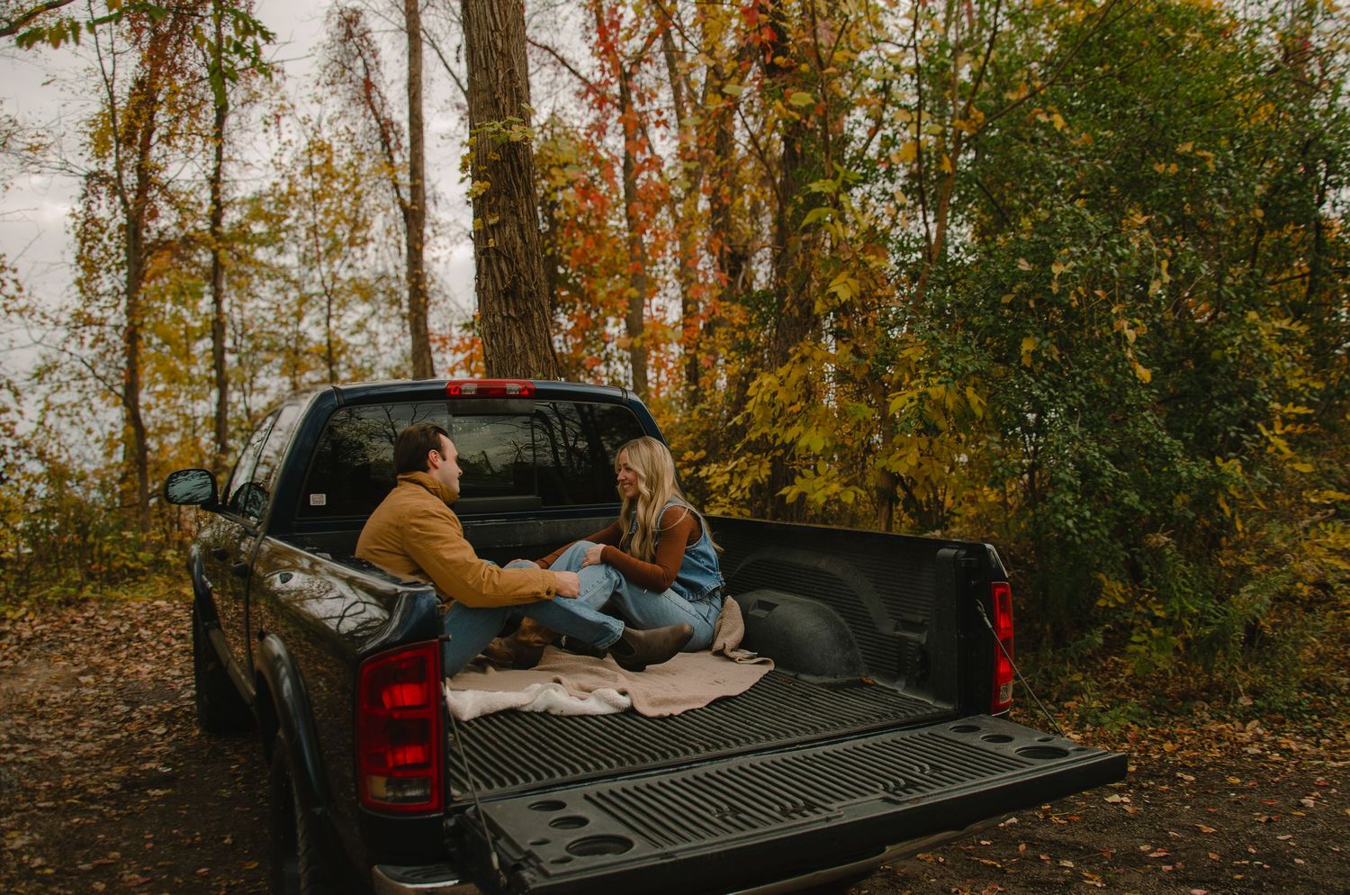 Couple relaxing in back of pickup truck with blankets surrounded by autumn trees during golden hour.