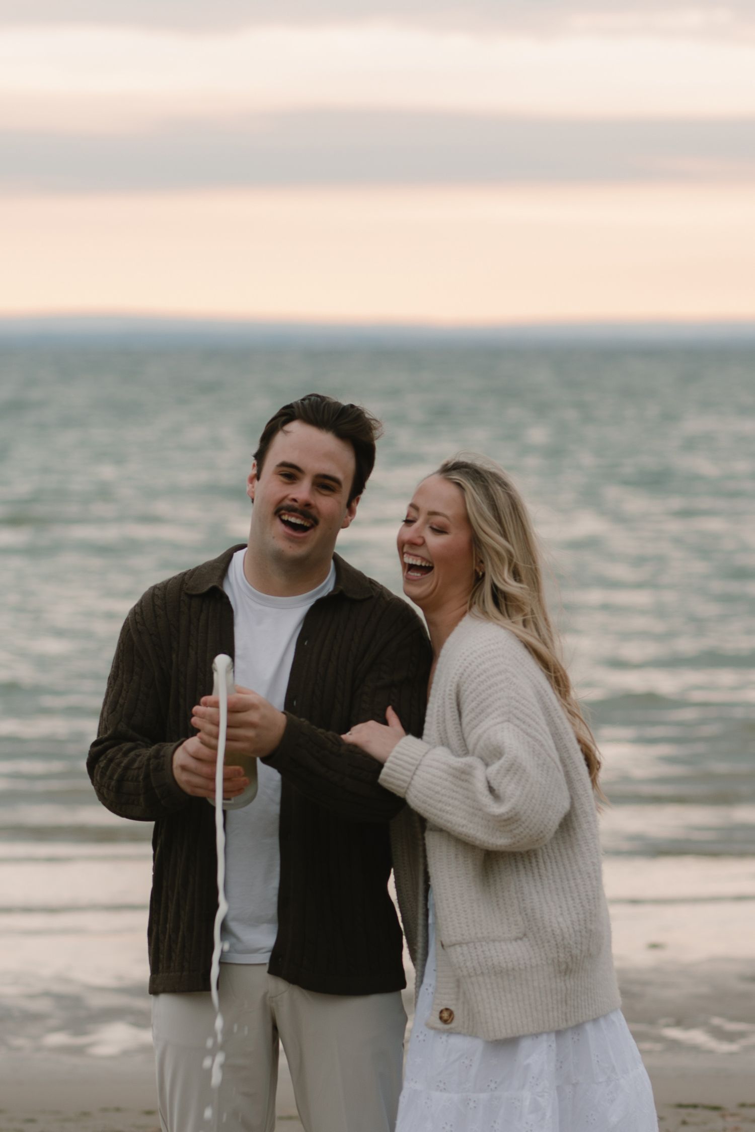 Happy couple laughs together while celebrating their engagement on a beach at dusk.