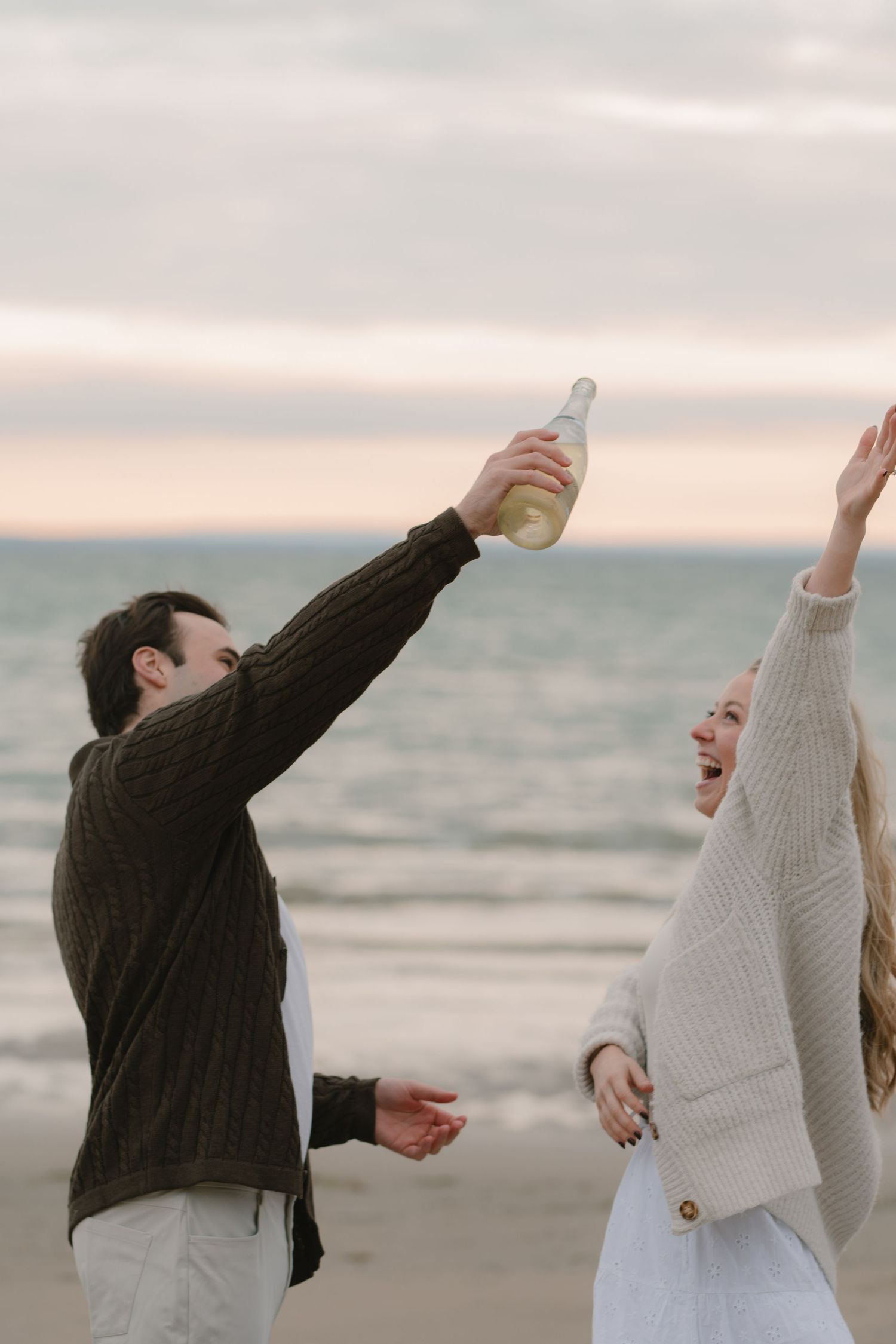 Couple in sweaters dancing playfully on beach with arms raised against ocean backdrop.