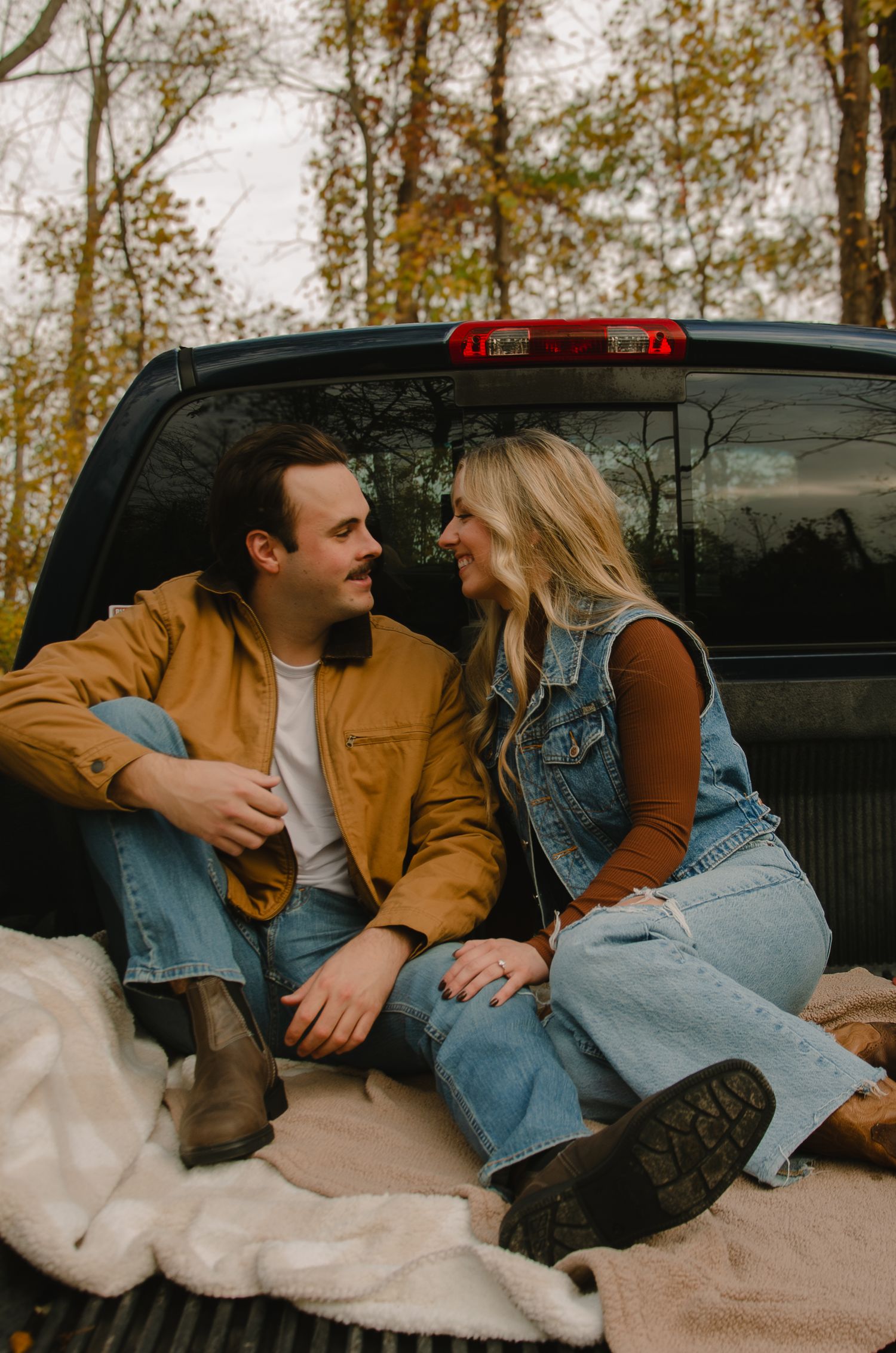 A couple sits together in the back of a pickup truck on a cozy blanket surrounded by autumn trees.