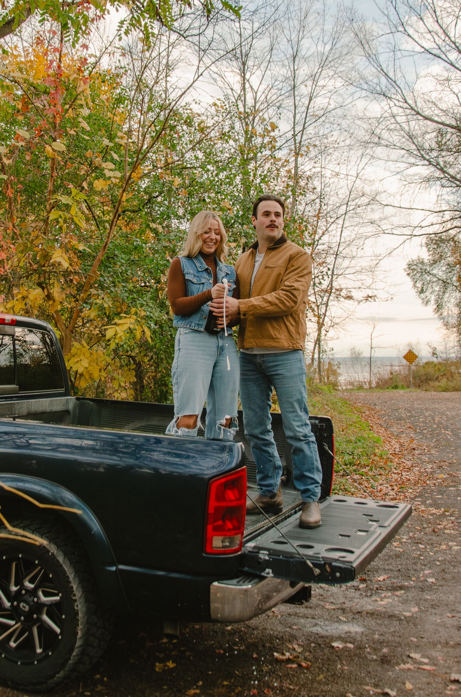 A couple sits on the tailgate of a black pickup truck surrounded by autumn foliage on a scenic country road.