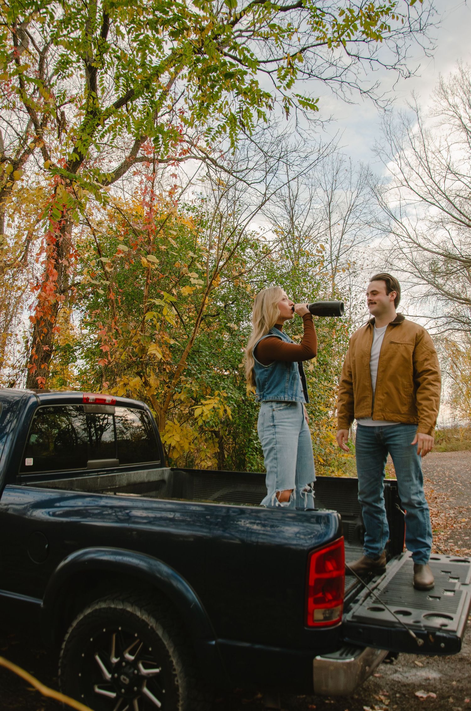 A couple in casual fall attire stands by a black pickup truck surrounded by autumn foliage.