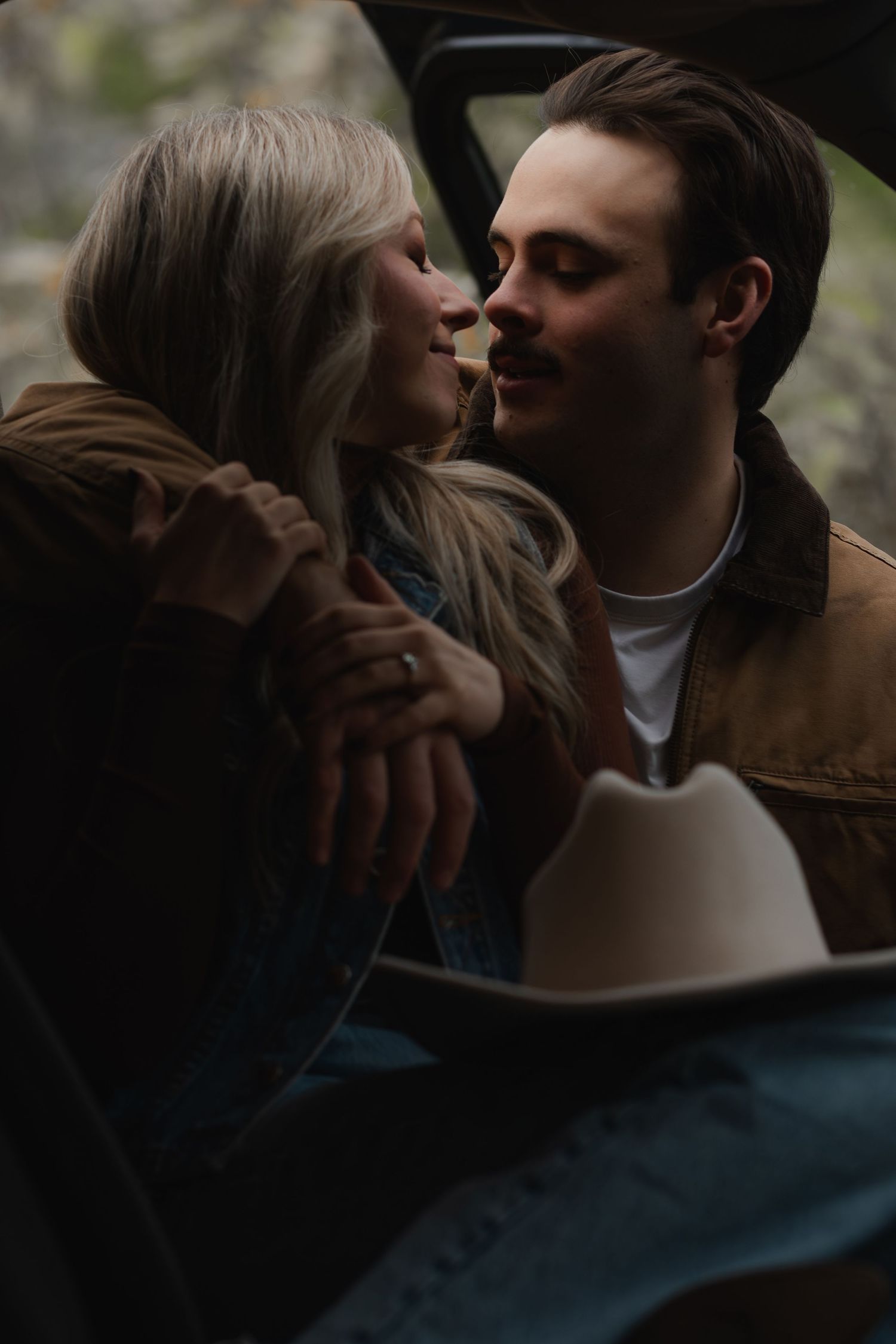 A romantic couple shares an intimate moment in a moody, dark setting with a white cowboy hat visible in the frame.