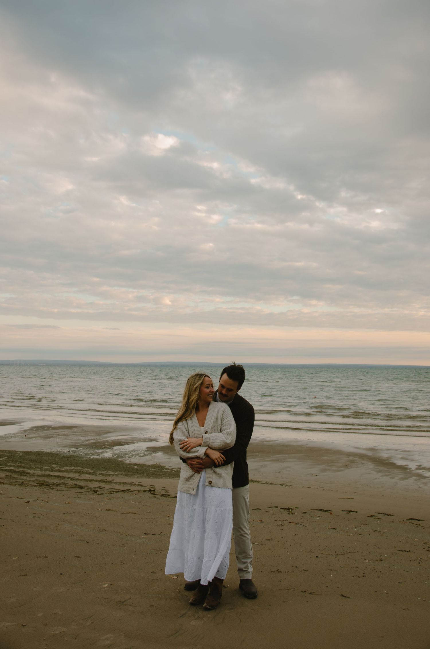 A romantic couple embracing on a cloudy beach at sunset.