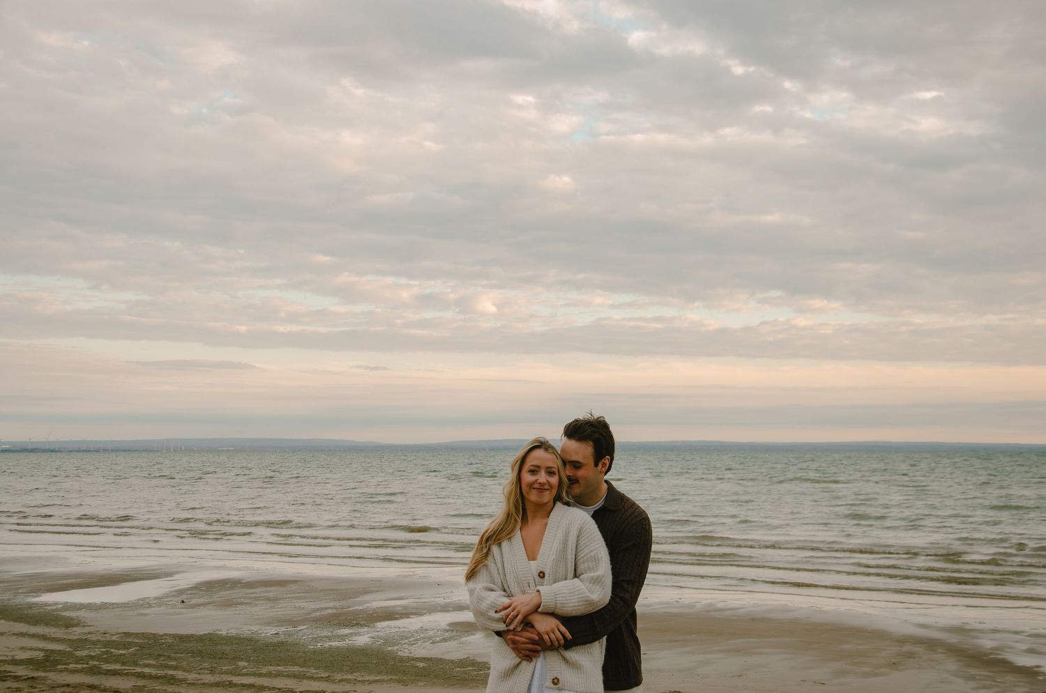A couple embraces on a moody beach at sunset with cloudy skies and waves rolling in along the shore.