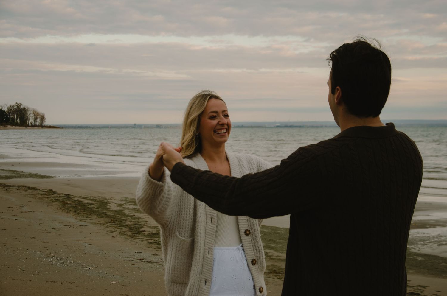 A couple walks along a cloudy beach shoreline at sunset.