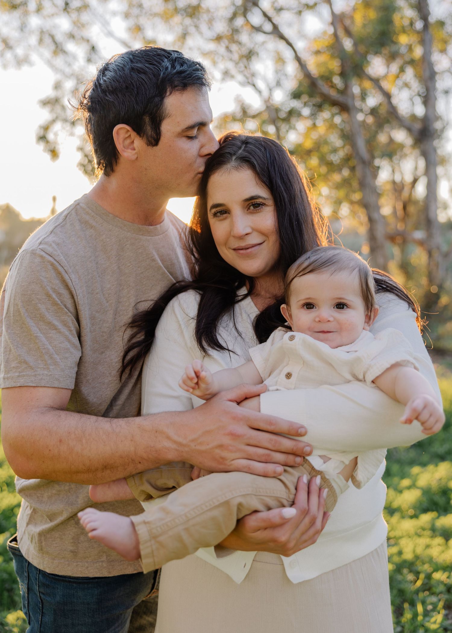 A family in coordinated sage green and white outfits poses together on a sandy beach at sunset.