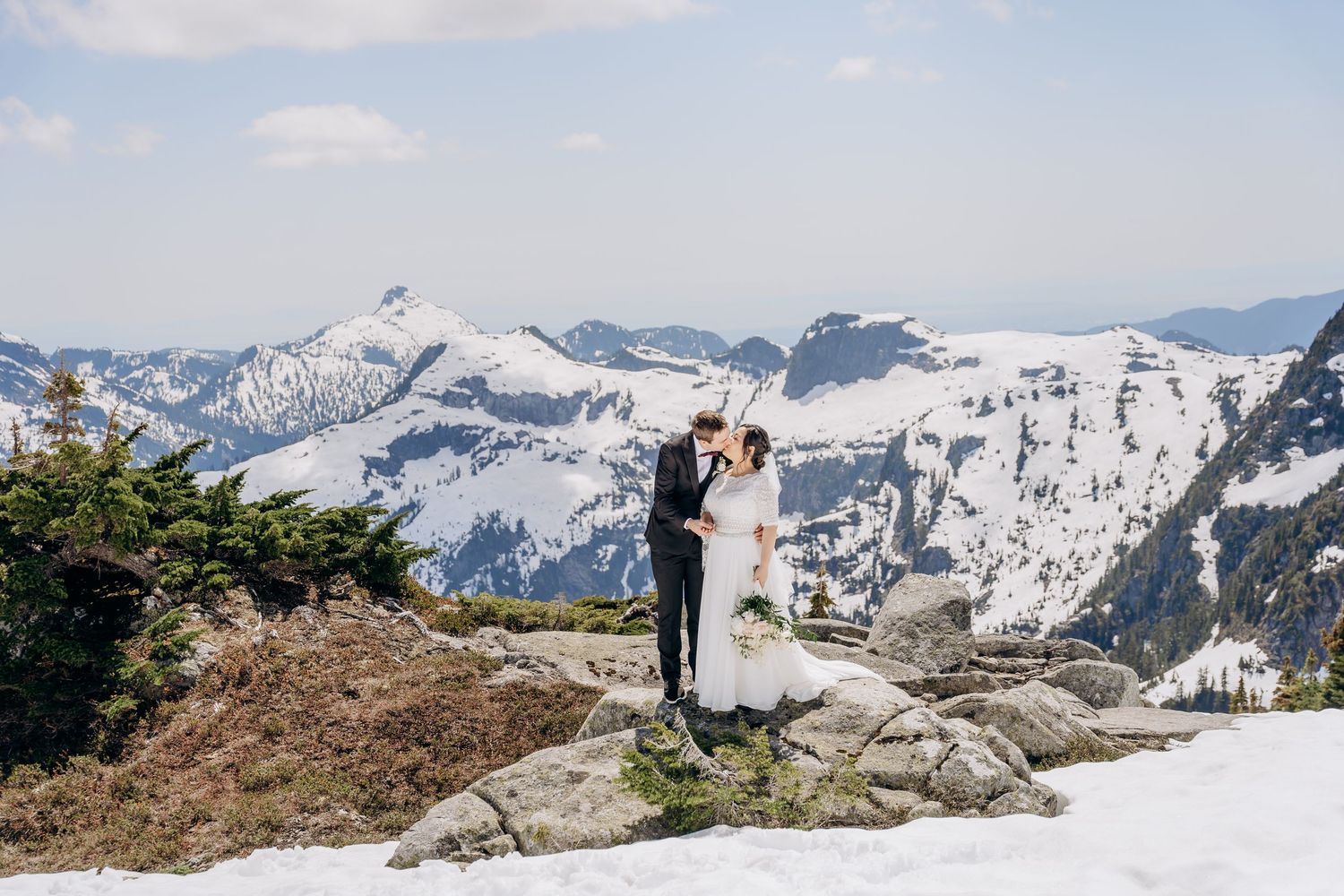 Panoramic view of snow-capped mountain peaks with hikers on a rocky ridge against a bright blue sky.