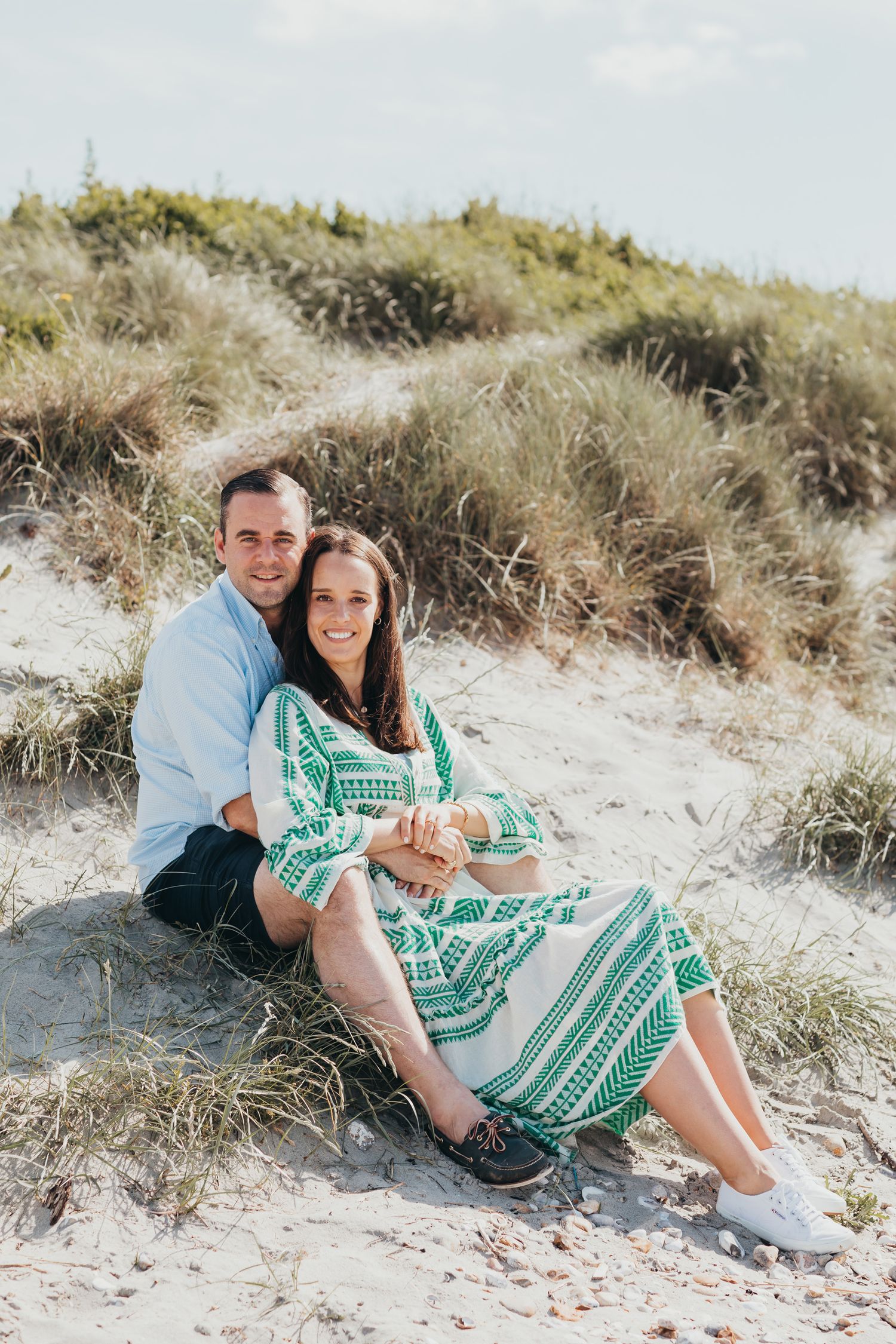 Romantic couple sits on sandy beach dunes, with woman wearing teal patterned dress.