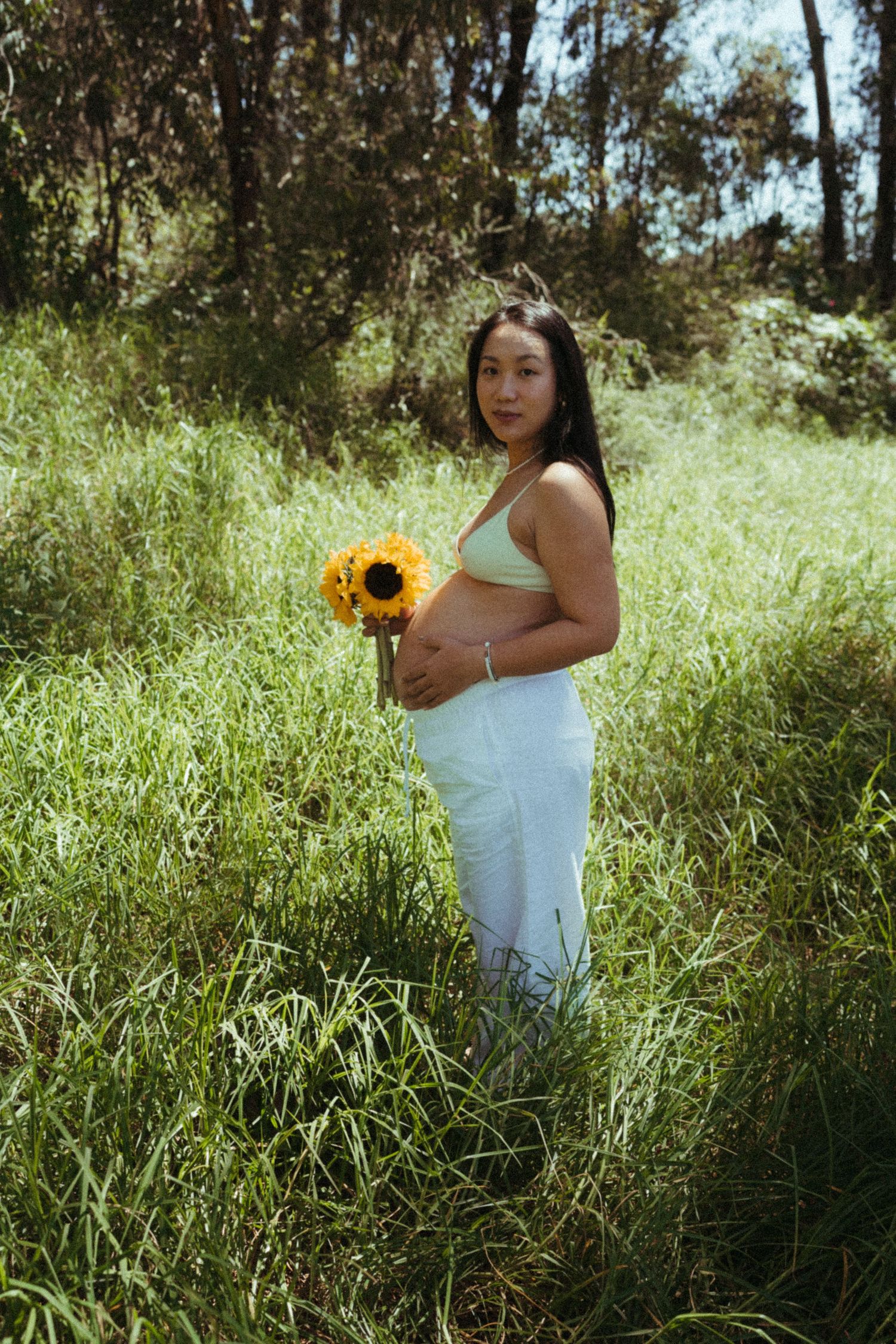A maternity photoshoot in a sunlit field with sunflowers and tall grass.