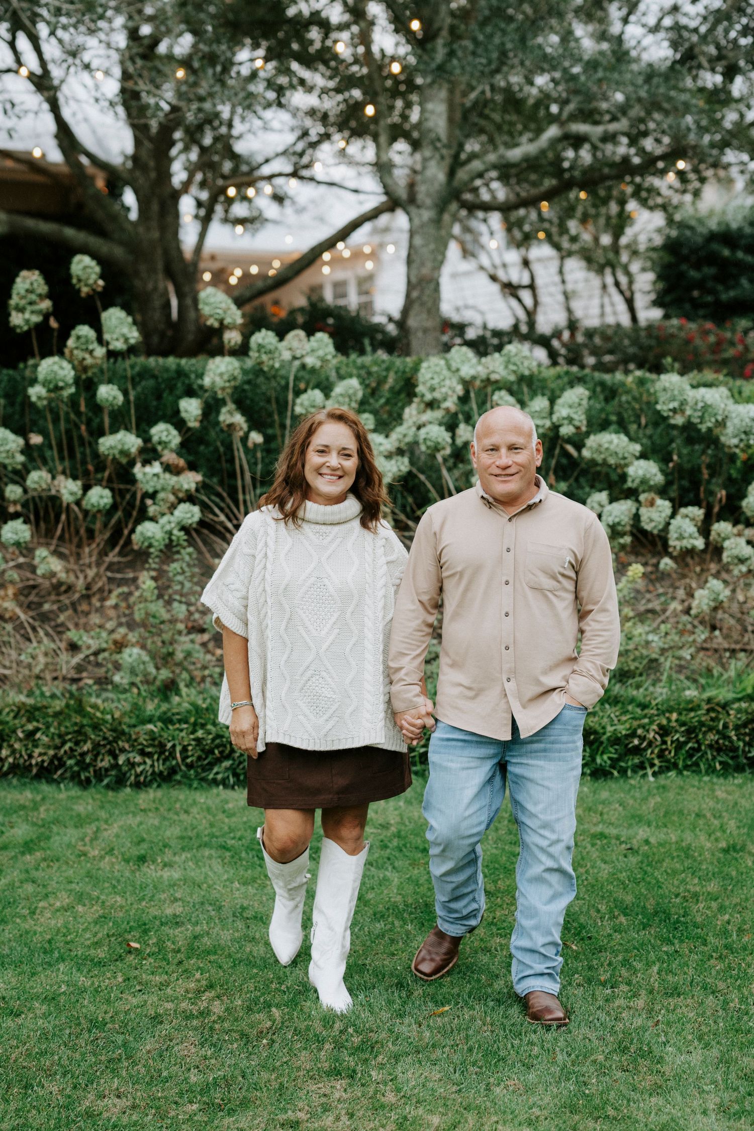 Two people walk together on green grass with white hydrangea flowers blooming in the background.