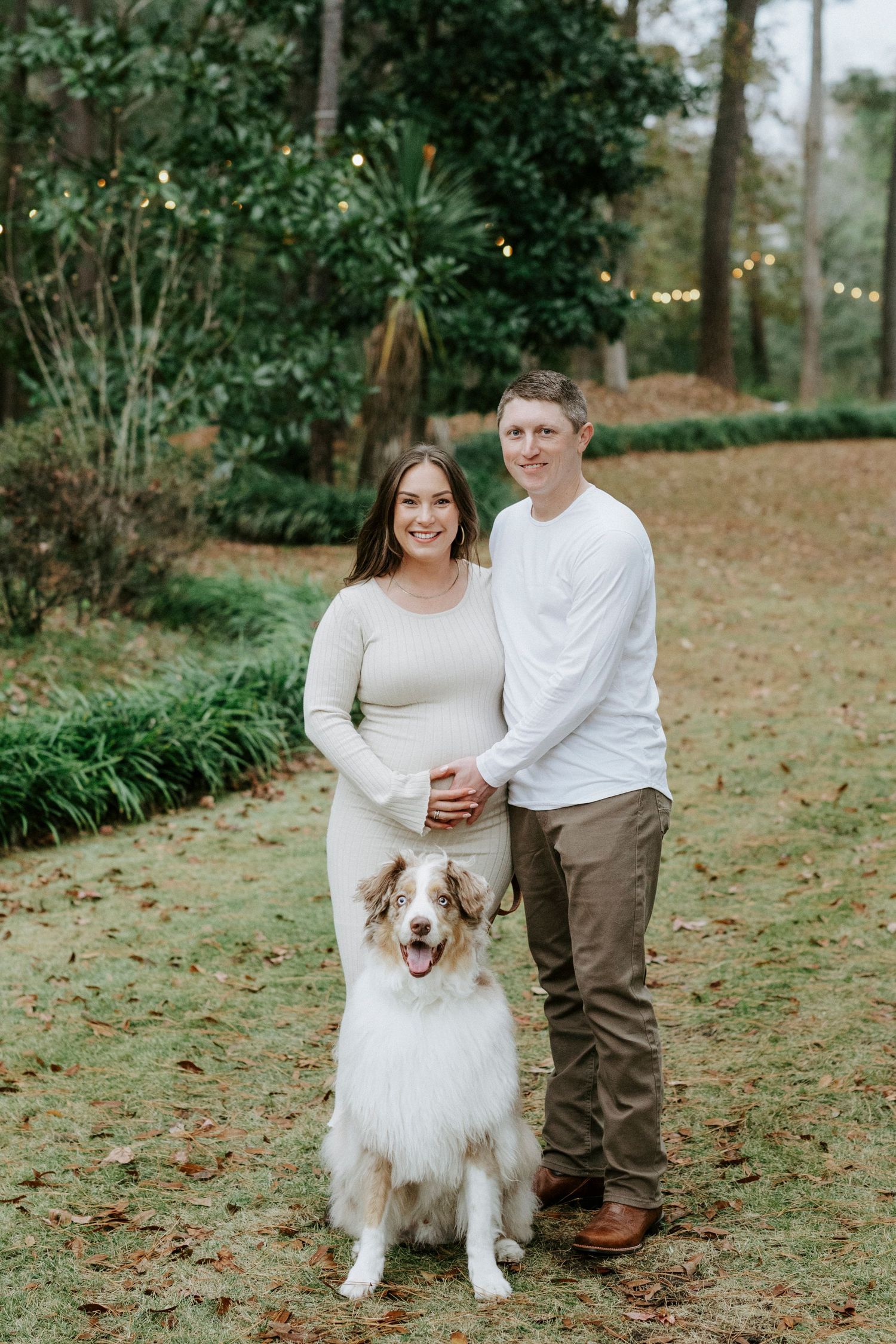 A couple in neutral-colored sweaters poses outdoors with their fluffy Australian Shepherd dog on autumn leaves.