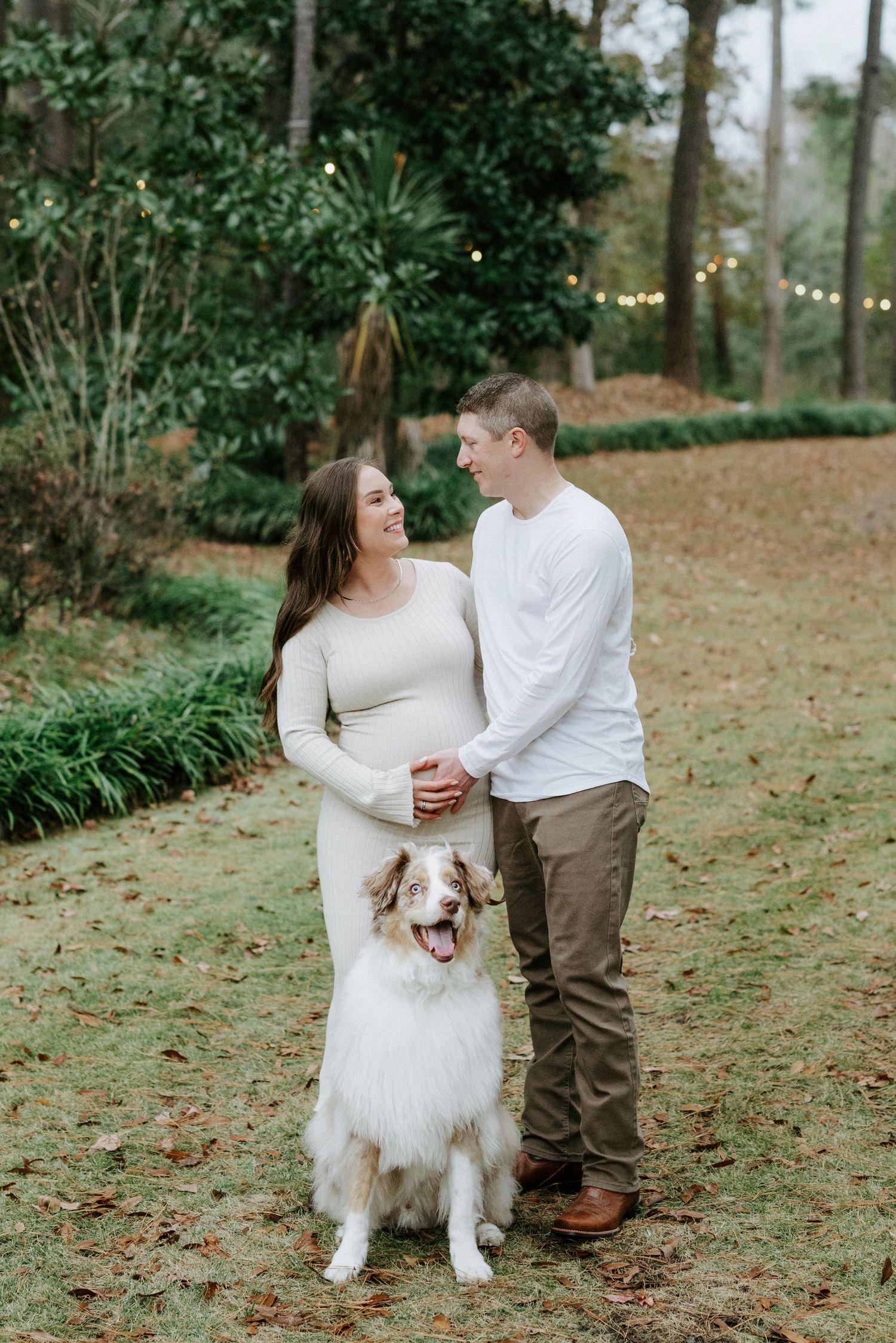 A couple in white and brown outfits poses outdoors with their fluffy white dog in a natural garden setting.