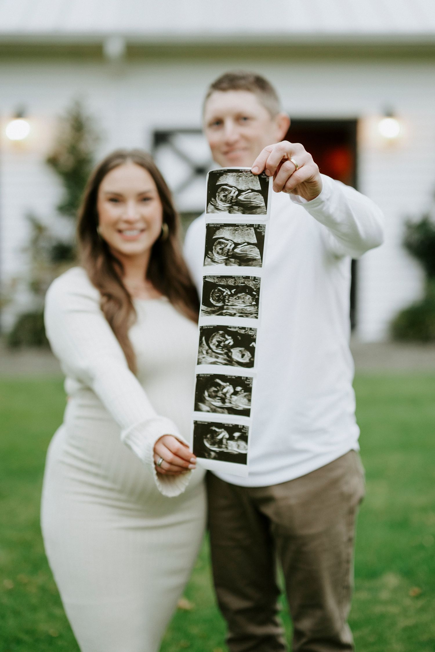 A couple in white and brown attire poses outdoors while holding ultrasound images to announce their pregnancy.