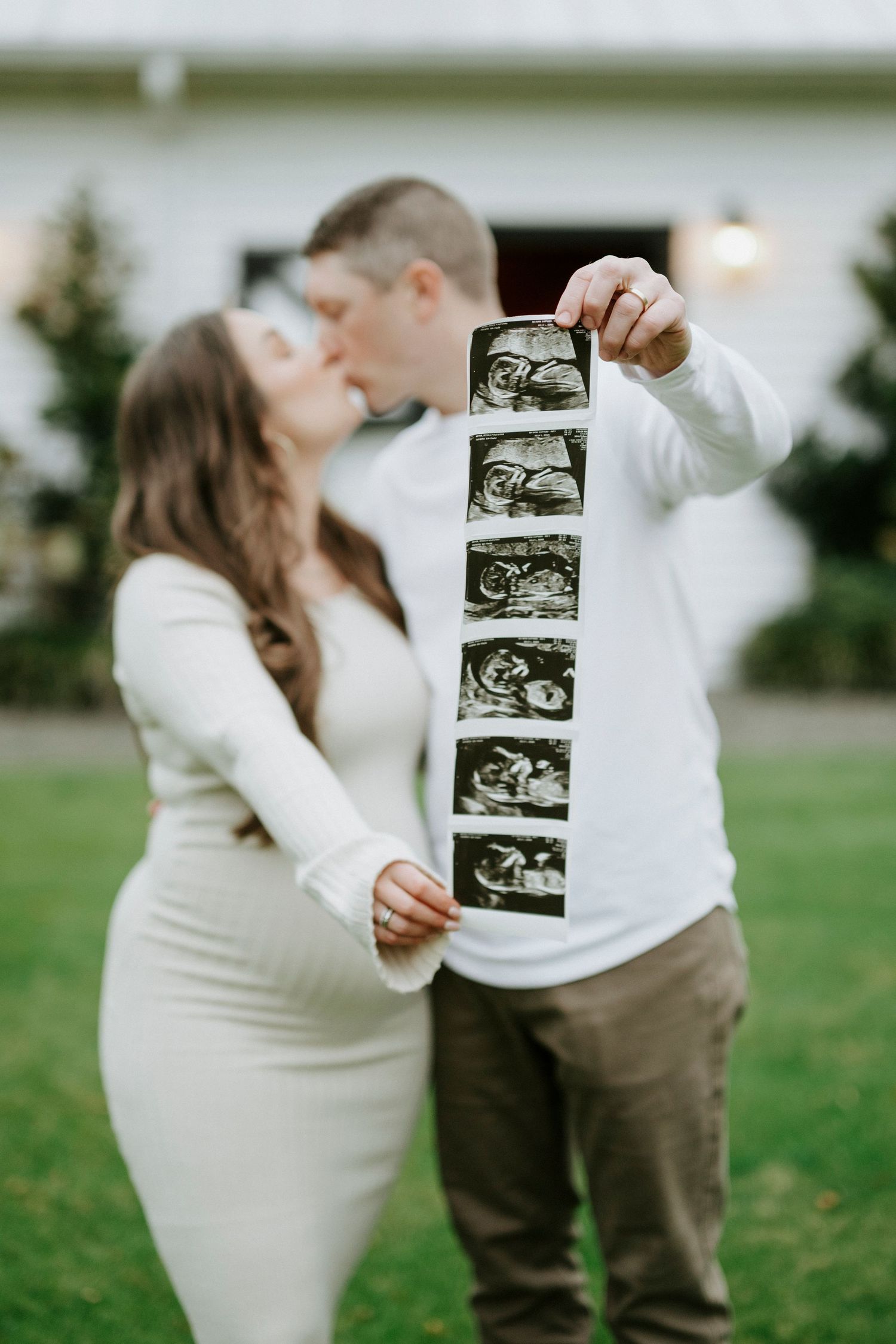 A couple in white sweaters holds up ultrasound photos while sharing an intimate moment on a grassy lawn.