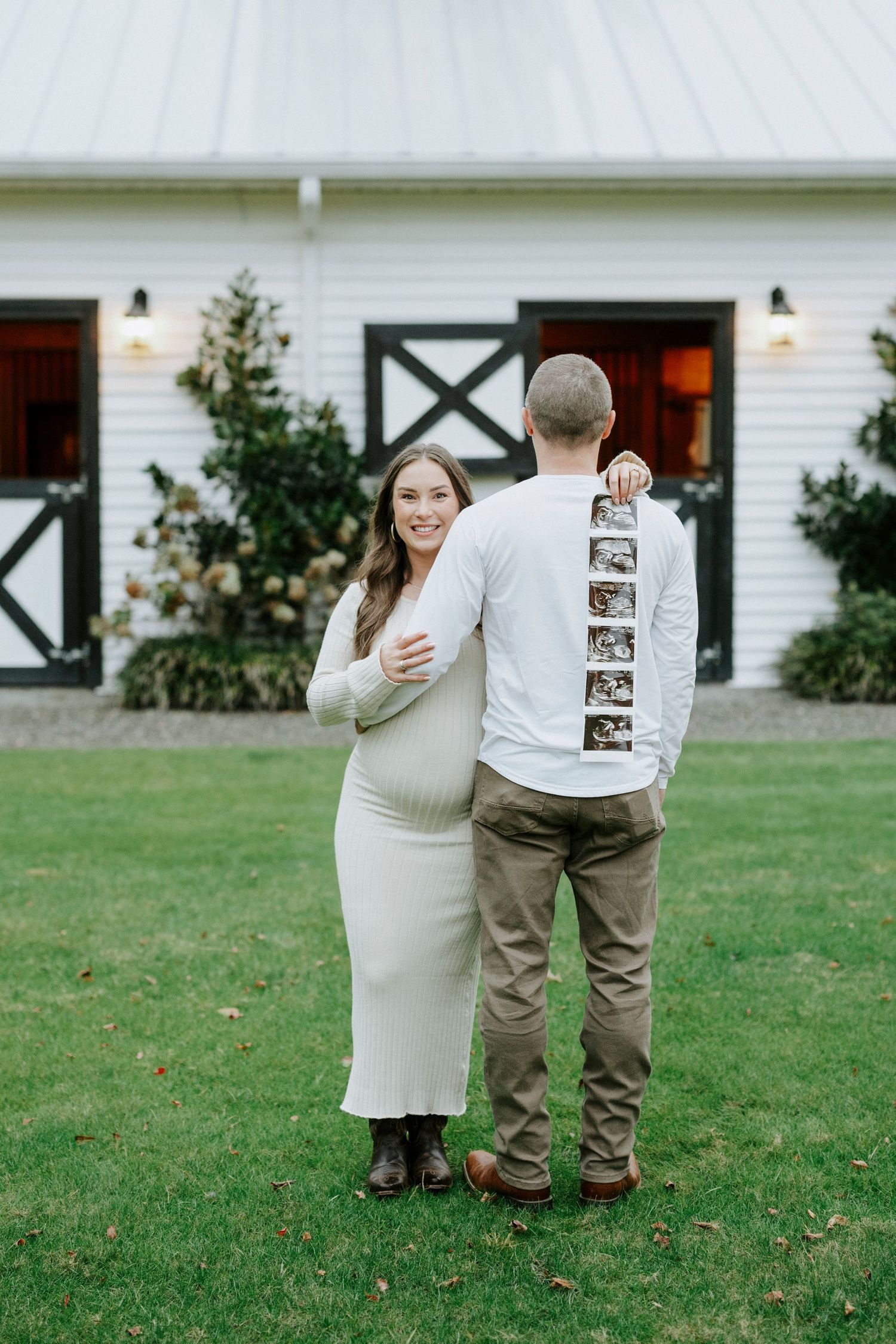 A couple poses in front of a white barn while displaying a strip of ultrasound photos on their backs.