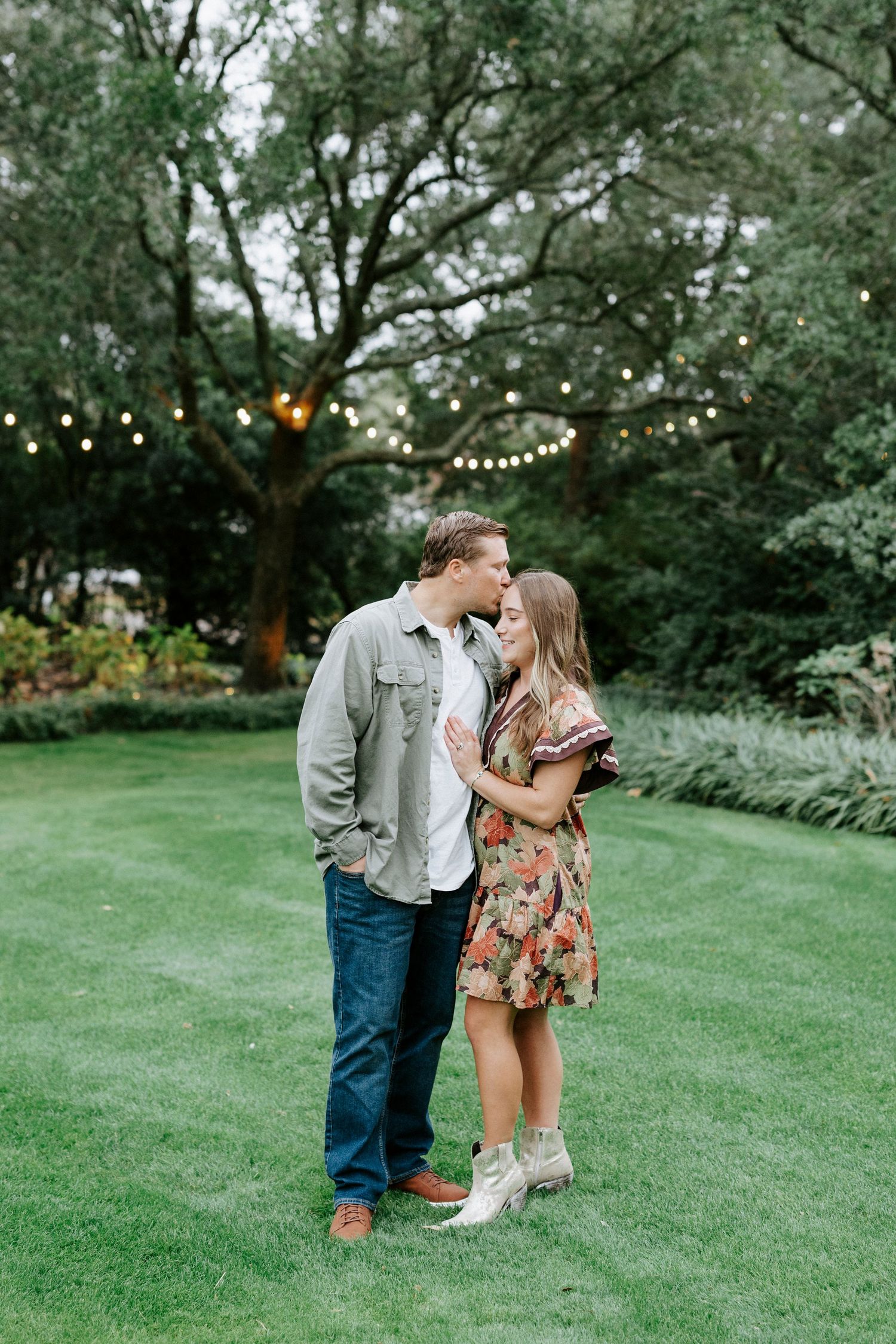 A romantic moment under string lights in a garden setting with trees in the background.