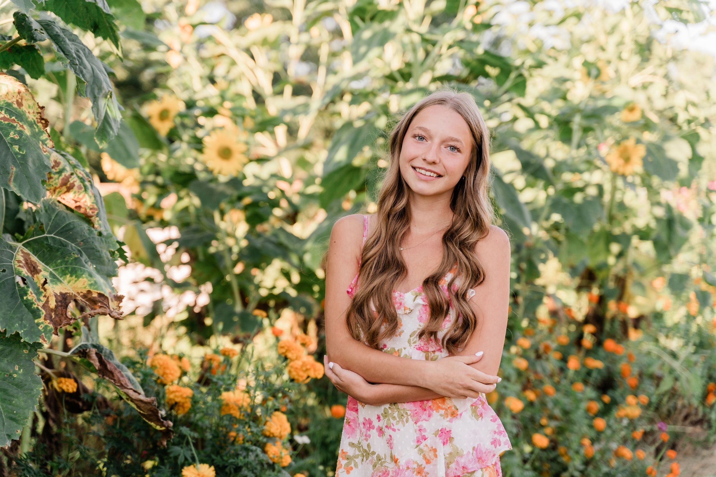 A person in a floral dress poses in a garden with blooming orange lantana flowers and green foliage.