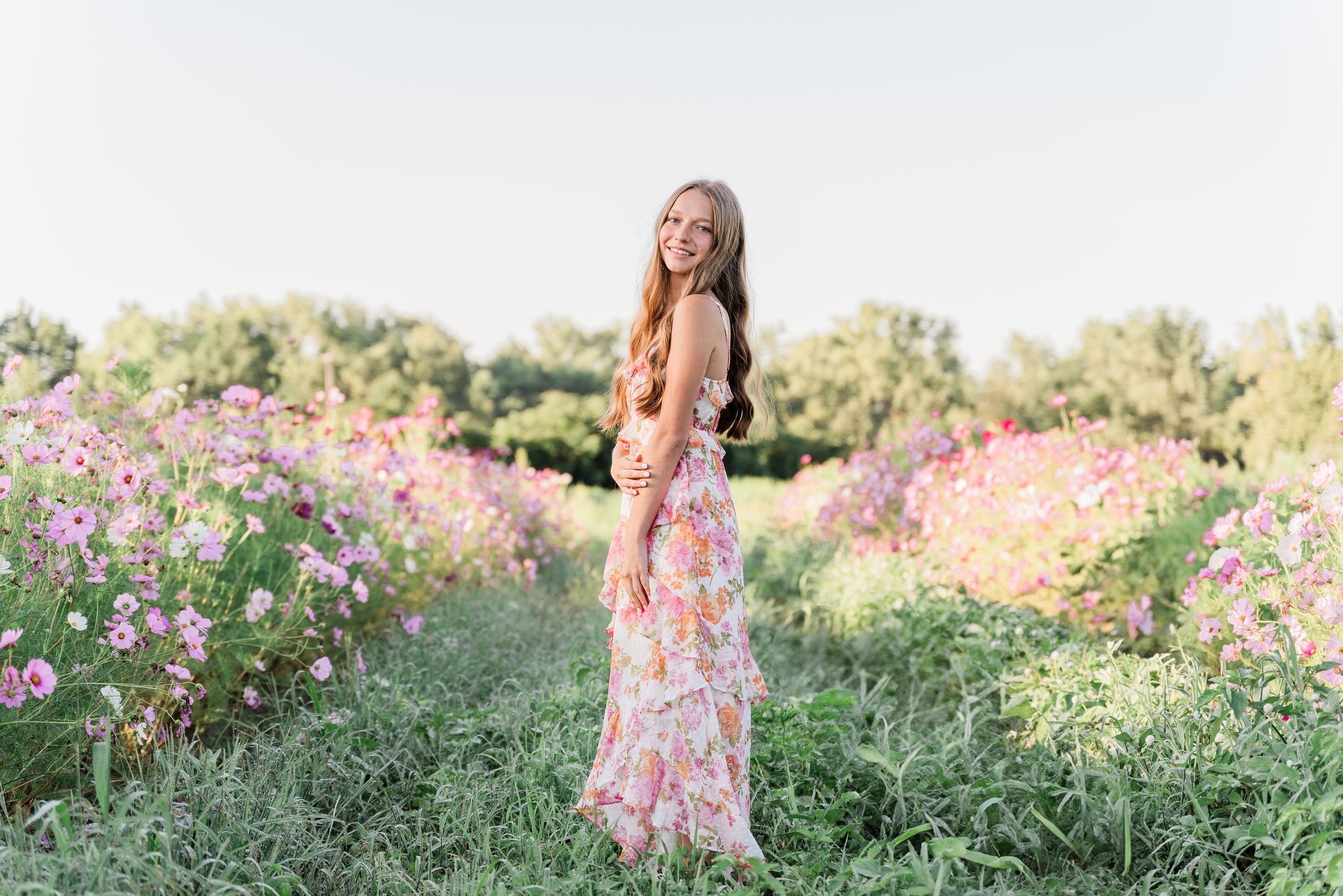 A figure in a floral dress stands gracefully among blooming pink cosmos flowers in a summer garden.