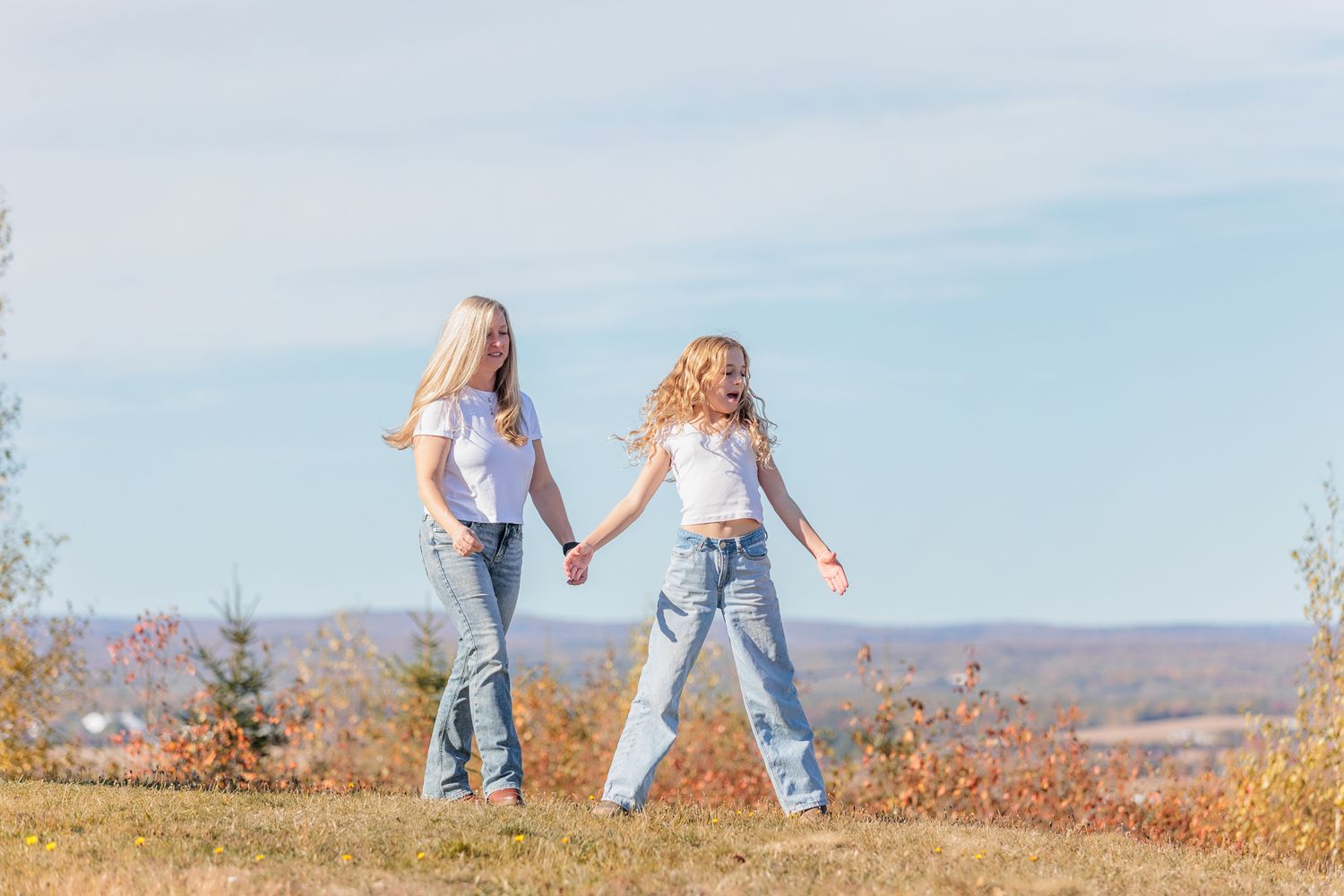 Two people in casual denim outfits holding hands while walking through an autumn field at sunset.