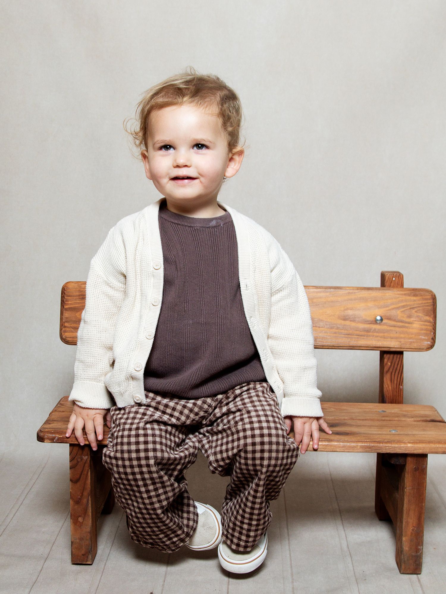 Studio shot of a little boy sitting on a bench smiling .  The background is cream matching the style of the photography through this website