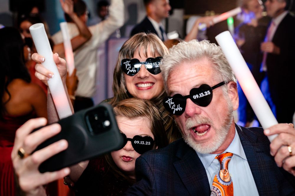 Wedding guests dance and celebrate with glowing LED foam light sticks on a crowded dance floor.
