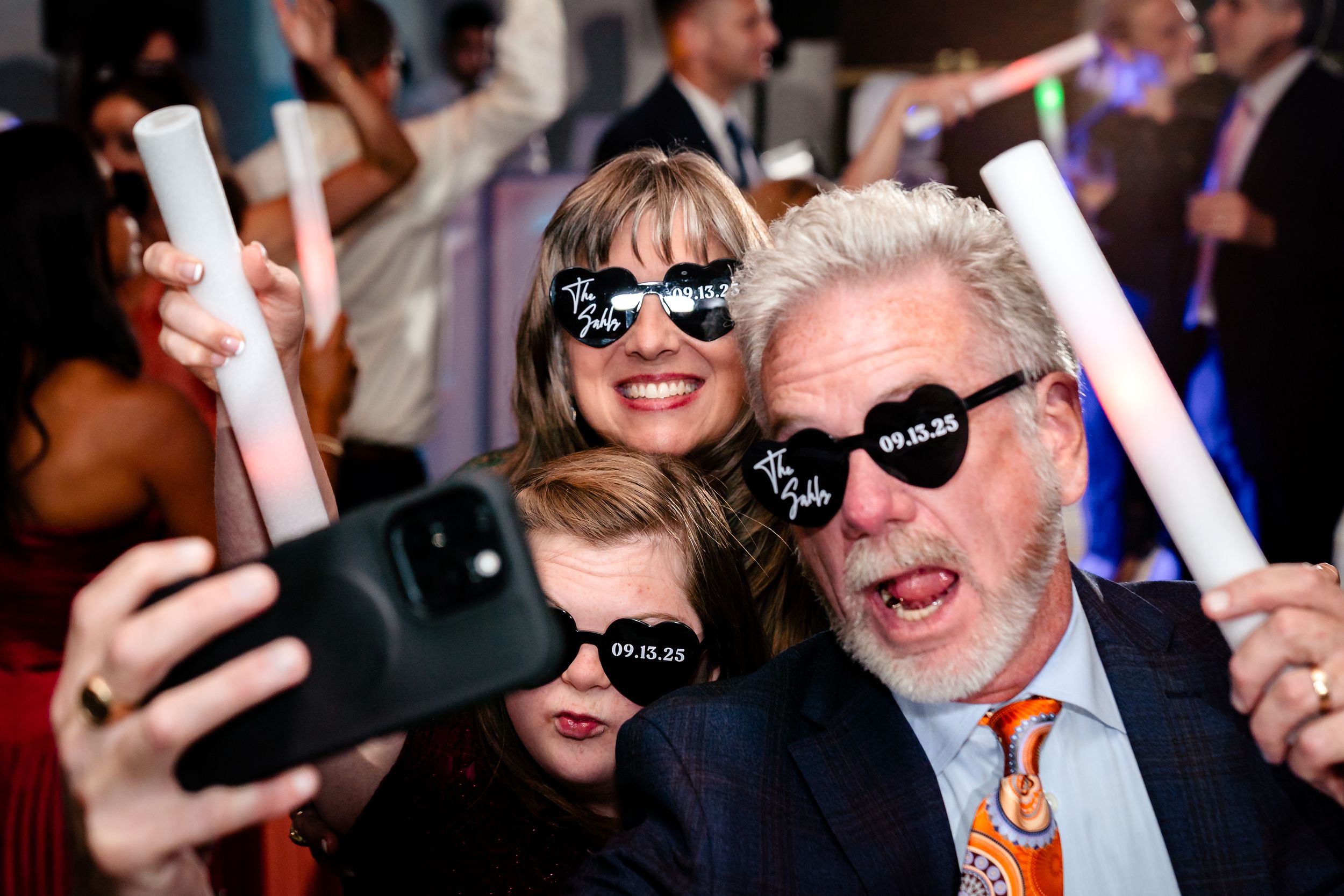 Wedding guests dance and celebrate with glowing LED foam light sticks on a crowded dance floor.