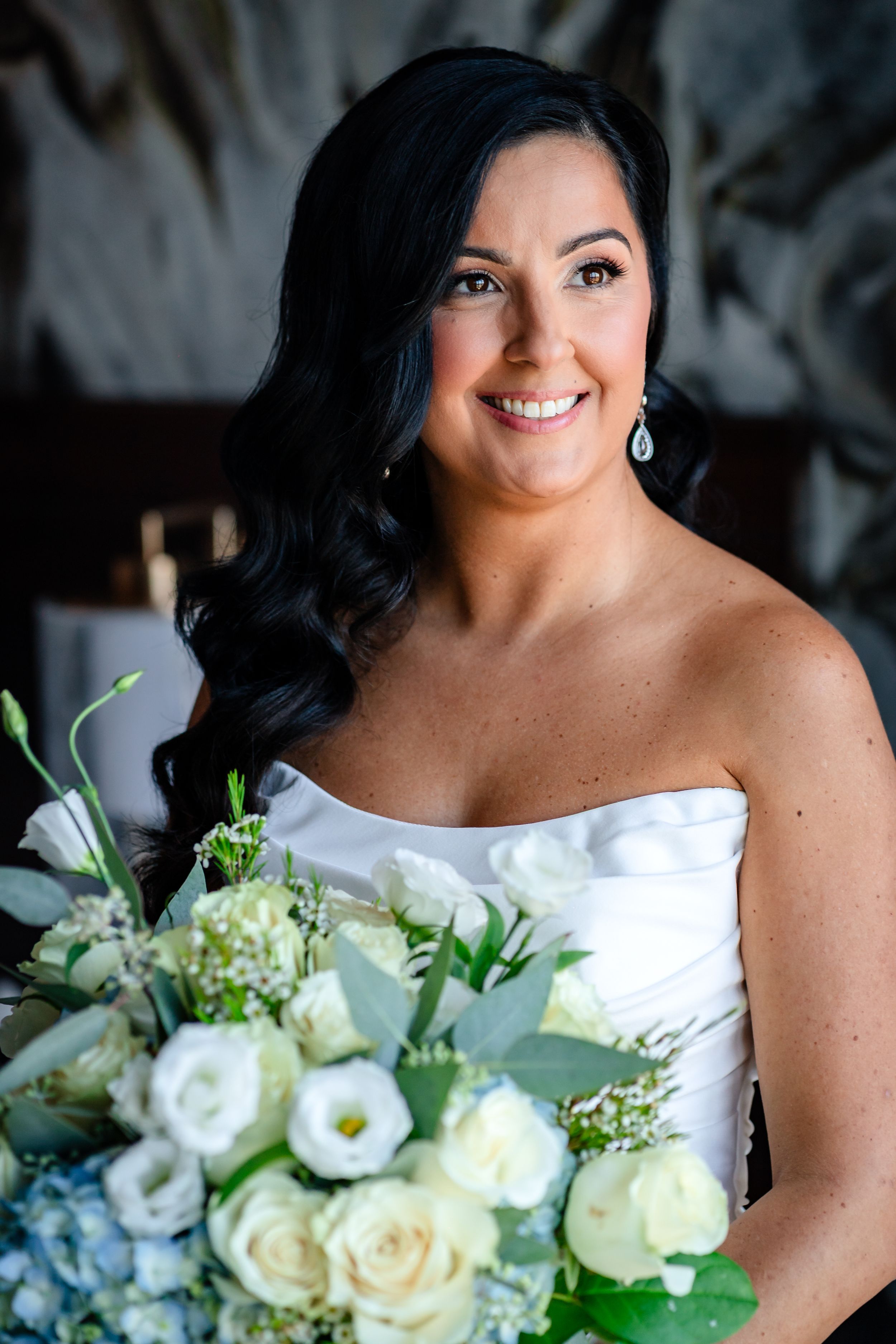 Elegant bridal portrait featuring a strapless white gown and fresh white and blue floral bouquet against dark background.