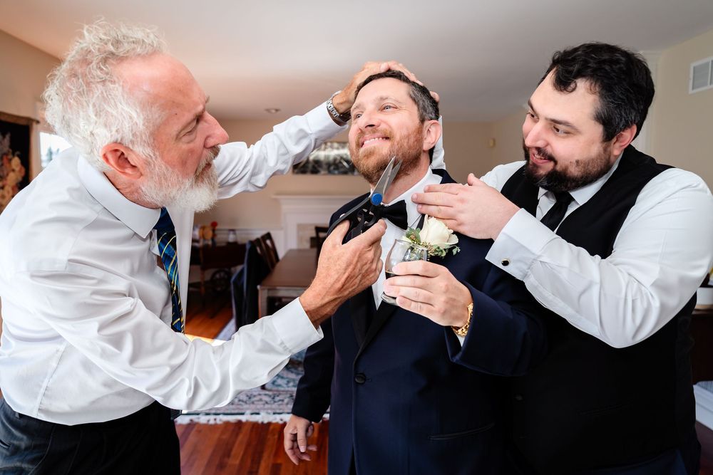 Three men help adjust a bow tie and groom each other while sharing laughs during wedding preparations.