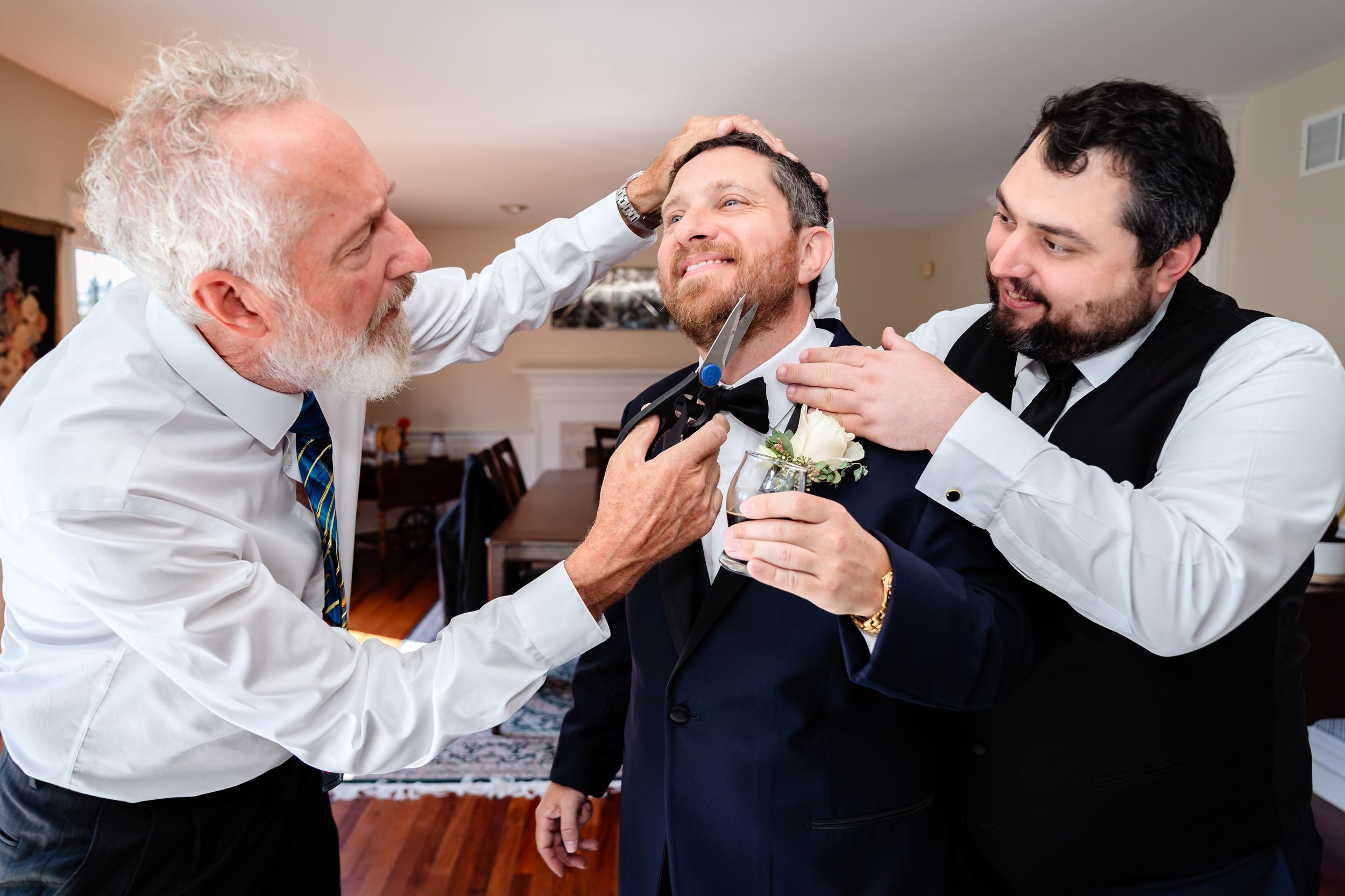 Three men help adjust a bow tie and groom each other while sharing laughs during wedding preparations.