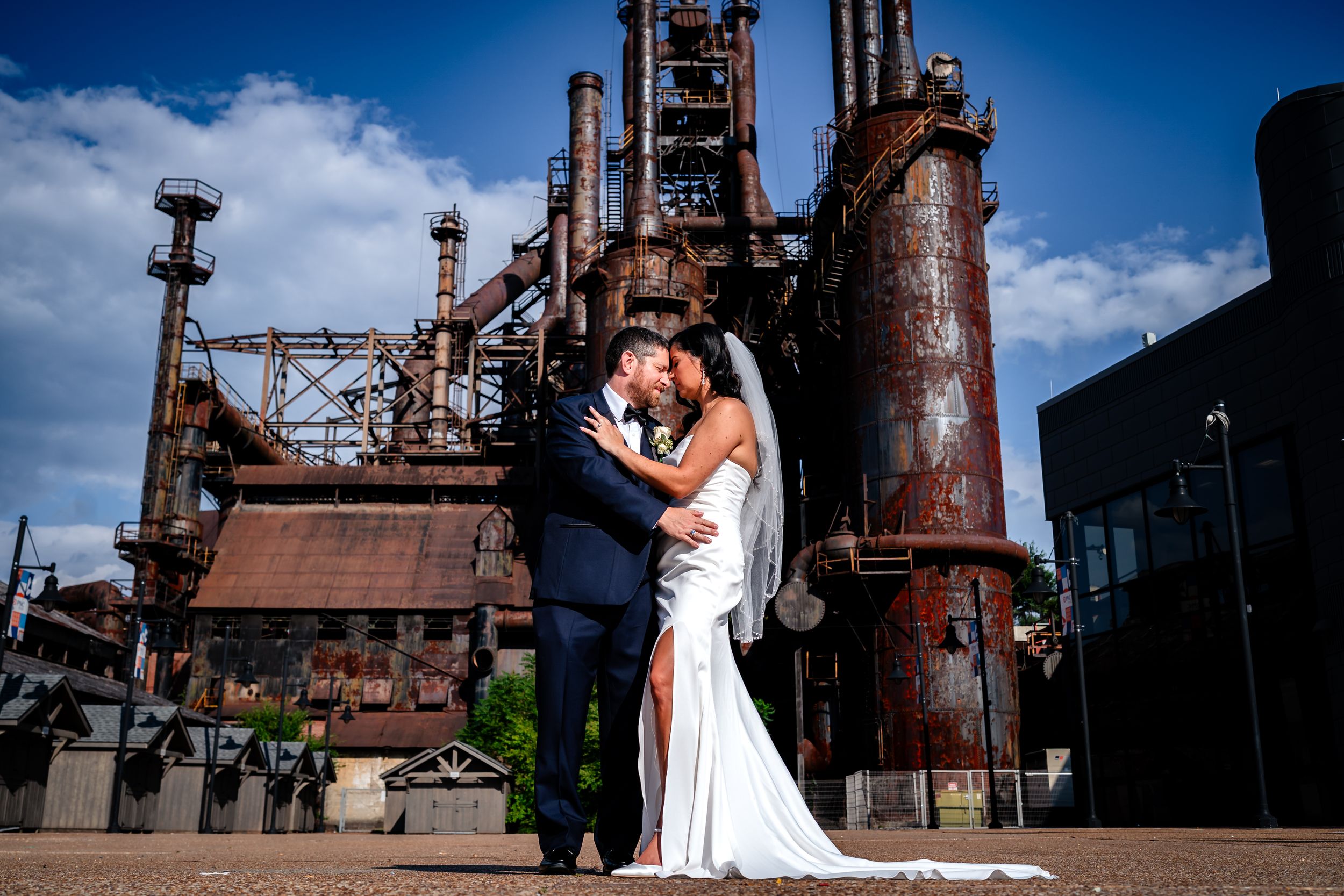 A couple in wedding attire embraces in front of rustic industrial steel mill architecture on their special day.