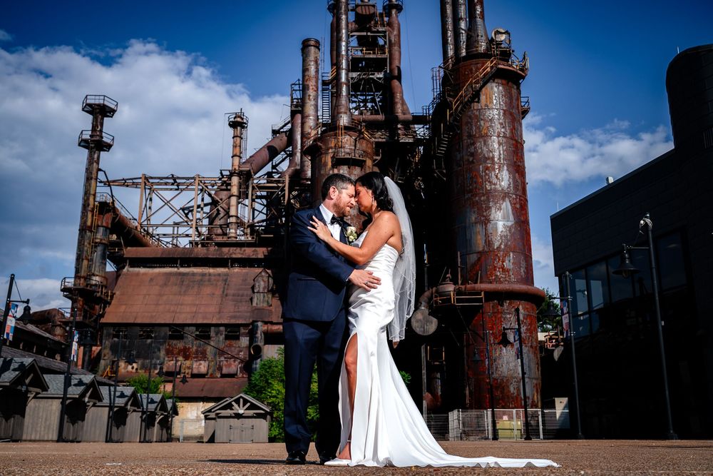 A couple in wedding attire embraces in front of rustic industrial steel mill architecture on their special day.
