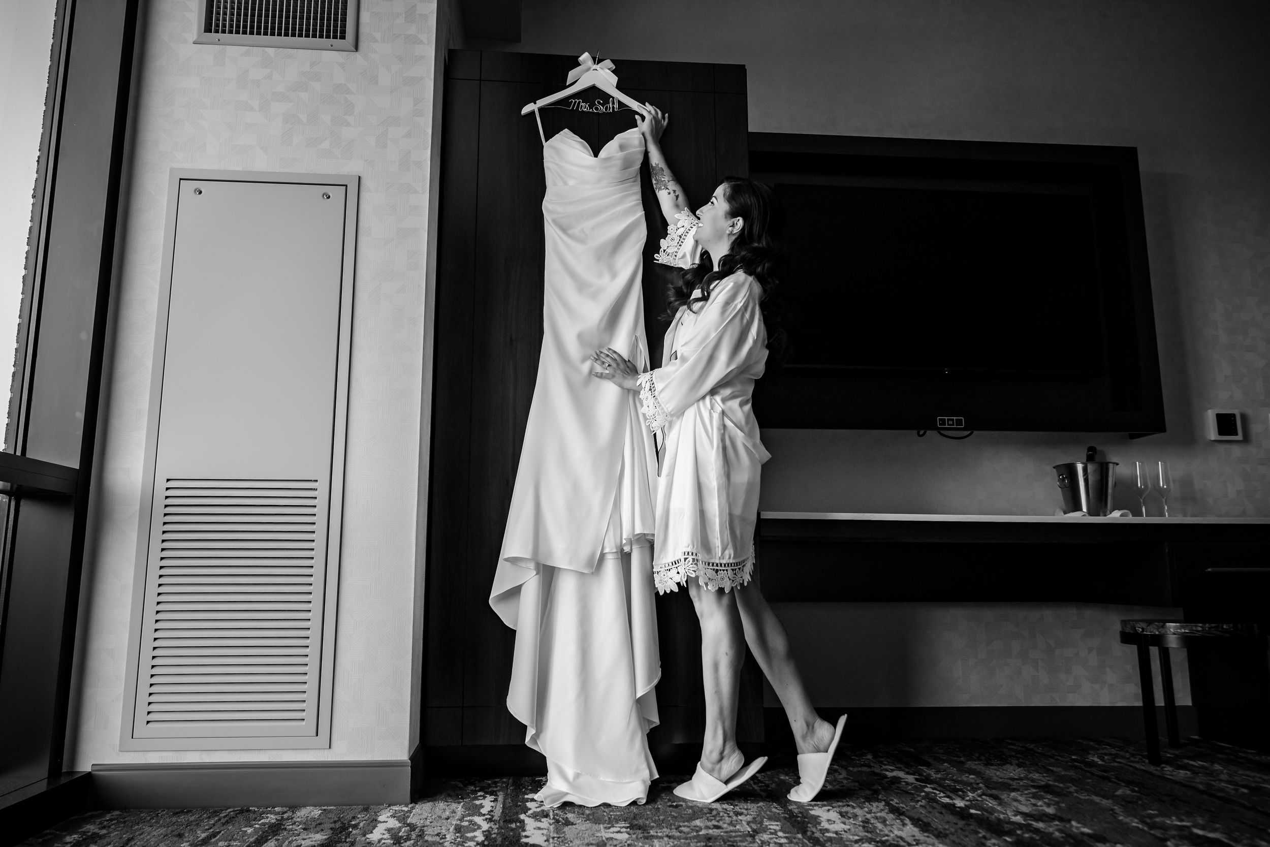 A wedding dress hangs on display as someone in a robe reaches up to touch it in a monochrome hotel room setting.