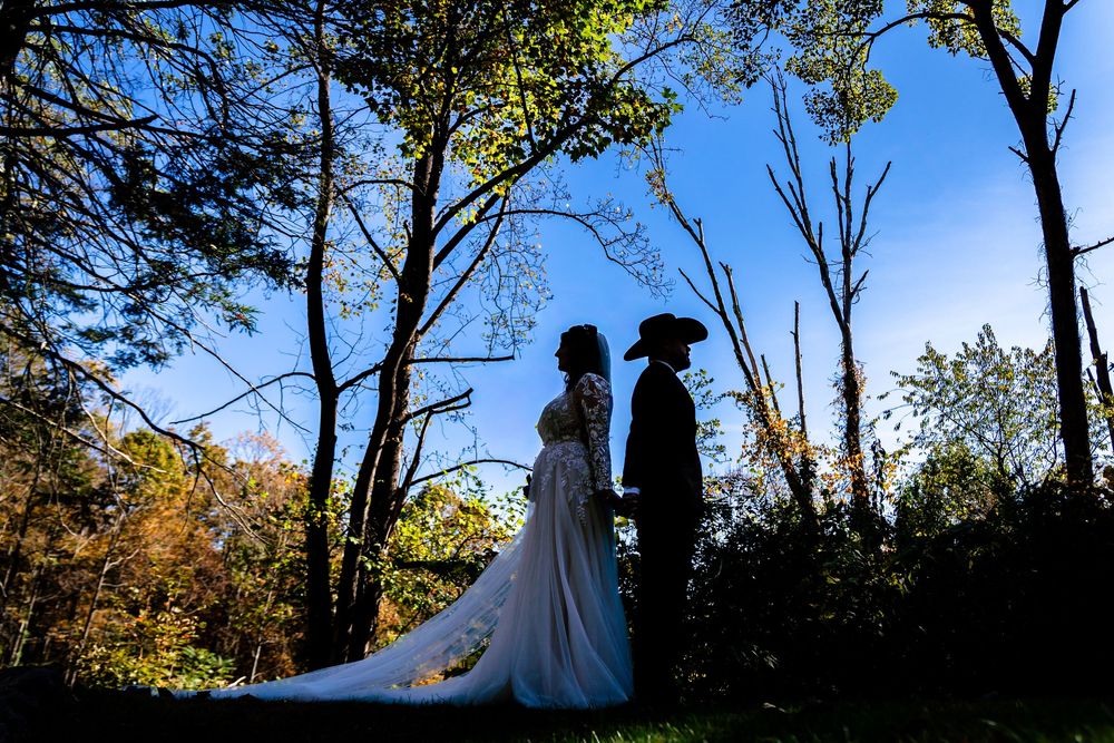 Silhouetted couple in wedding attire stands against dramatic sunset sky with trees in autumn forest setting.