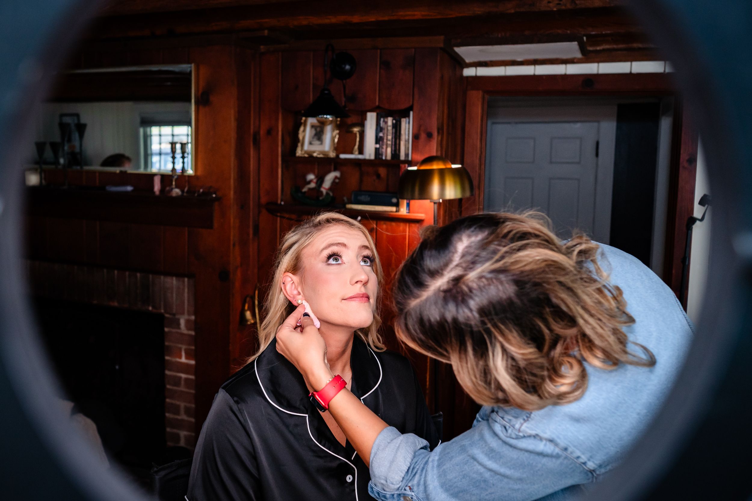 A makeup artist applies cosmetics to a model in a dimly lit room with wooden paneling.