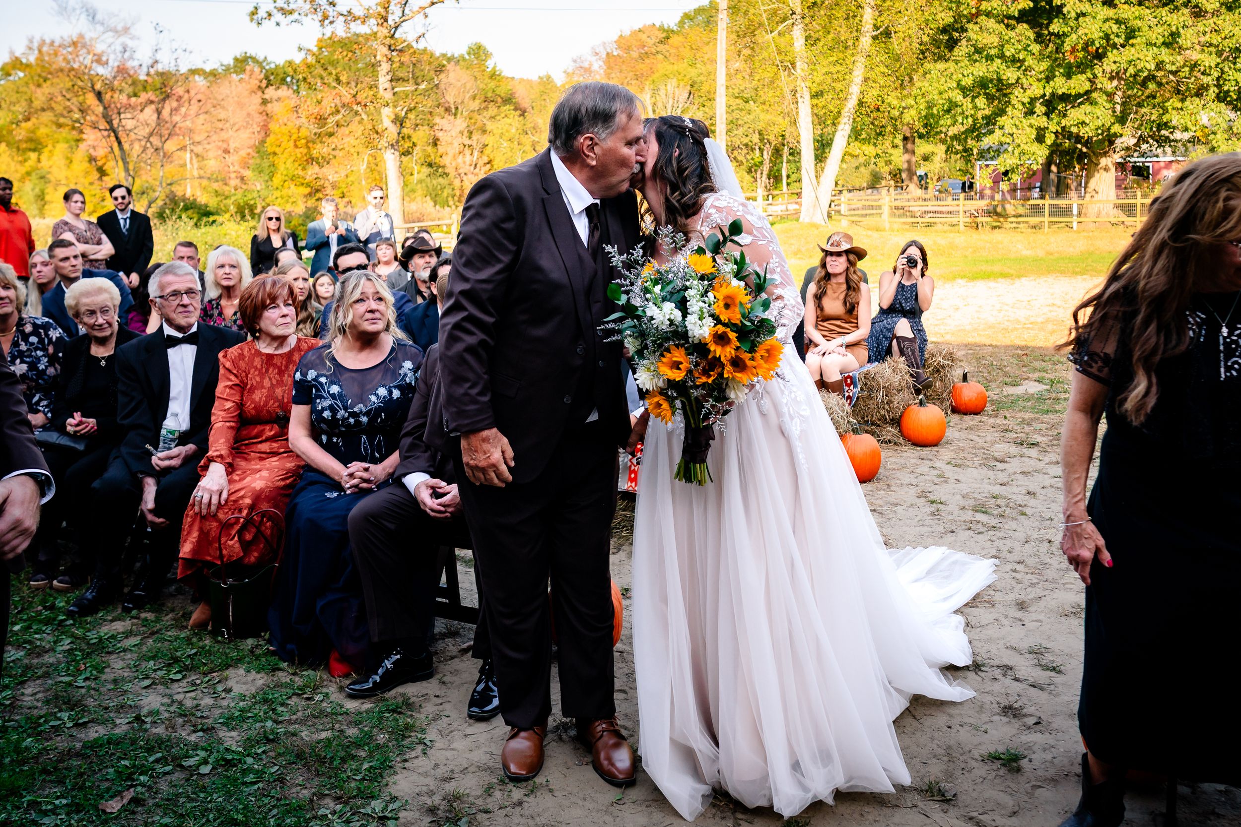 Wedding guests gather outdoors at sunset during a fall ceremony with autumn foliage and decorative pumpkins.