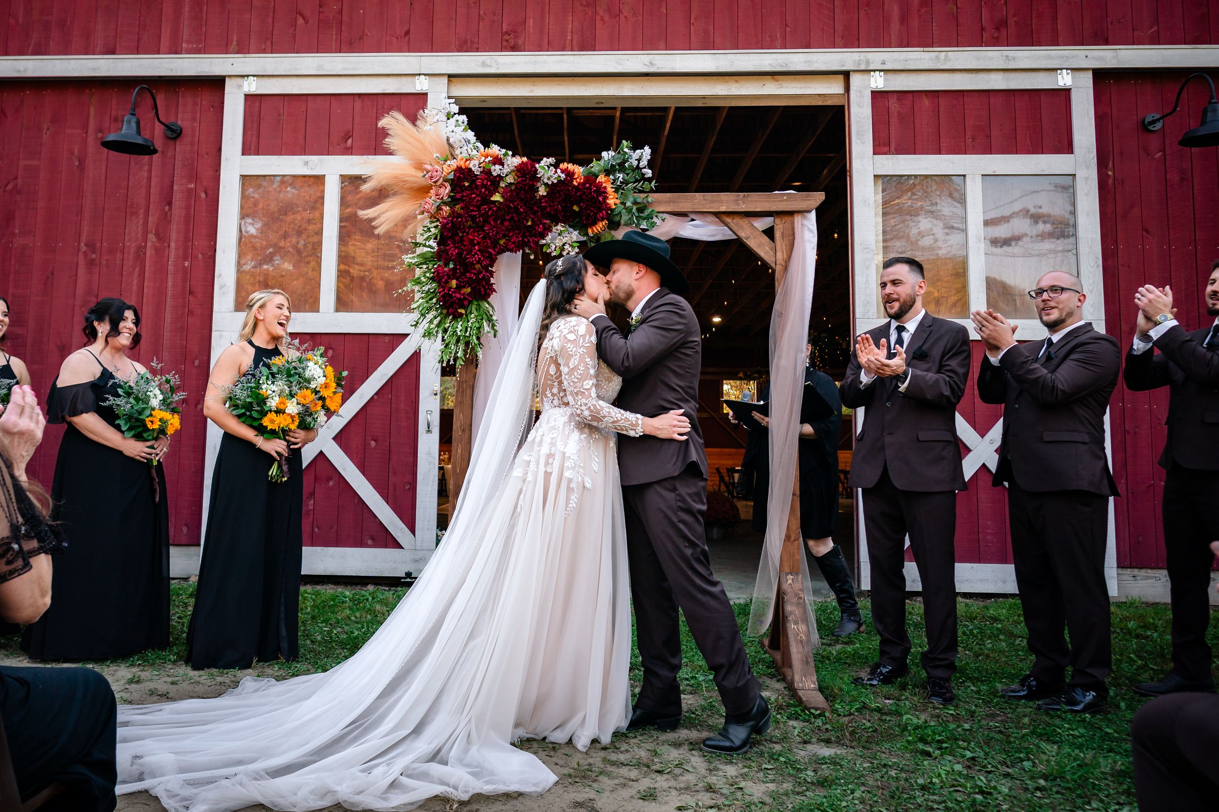 Wedding party members line up against a rustic red barn wall for formal photos during a countryside celebration.