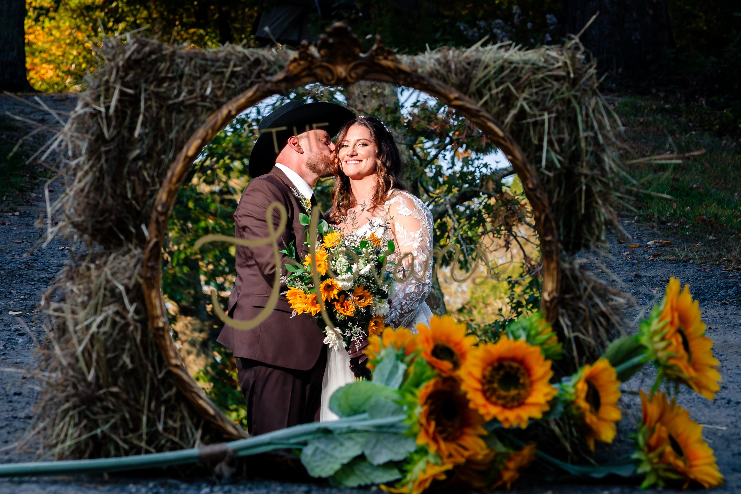 A rustic wedding scene framed by a wooden arch with bright sunflowers in the foreground during golden hour.