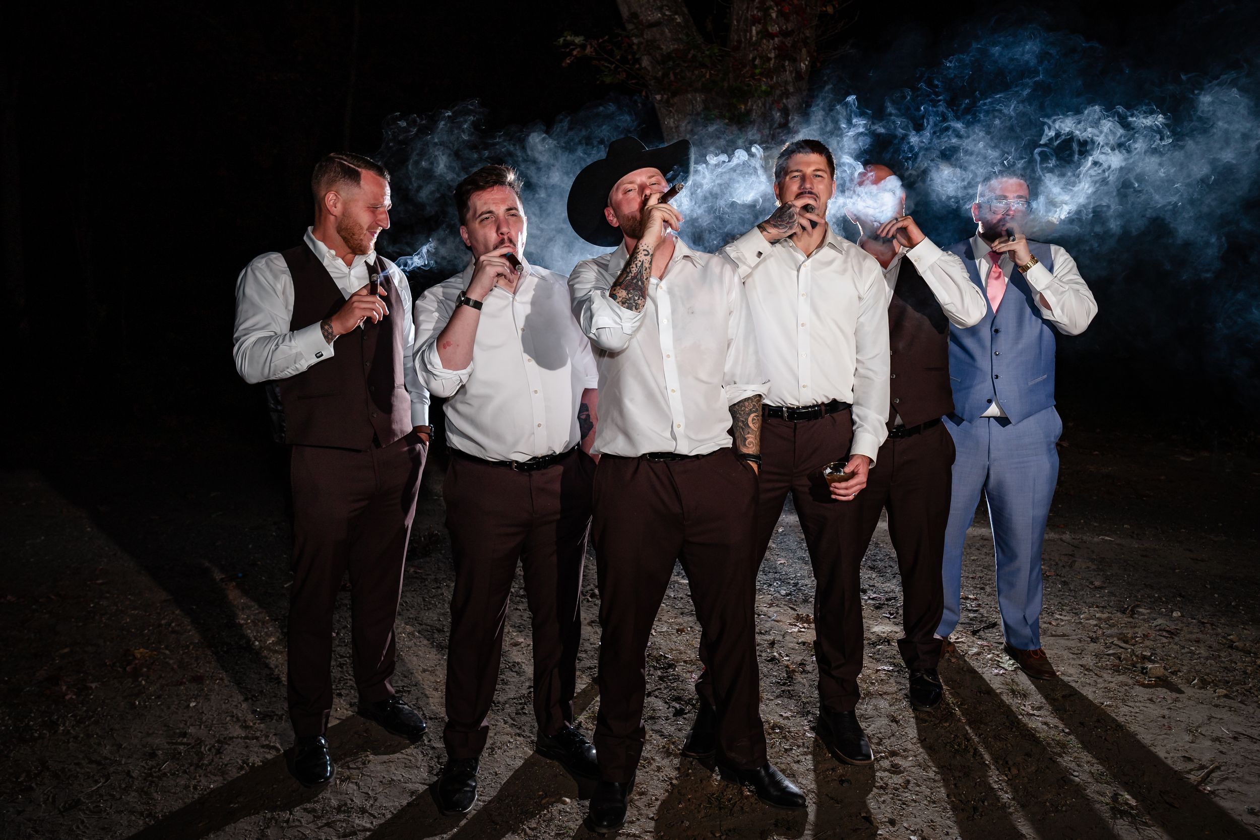 Groomsmen in western attire pose together while smoking cigars at an outdoor evening wedding celebration.
