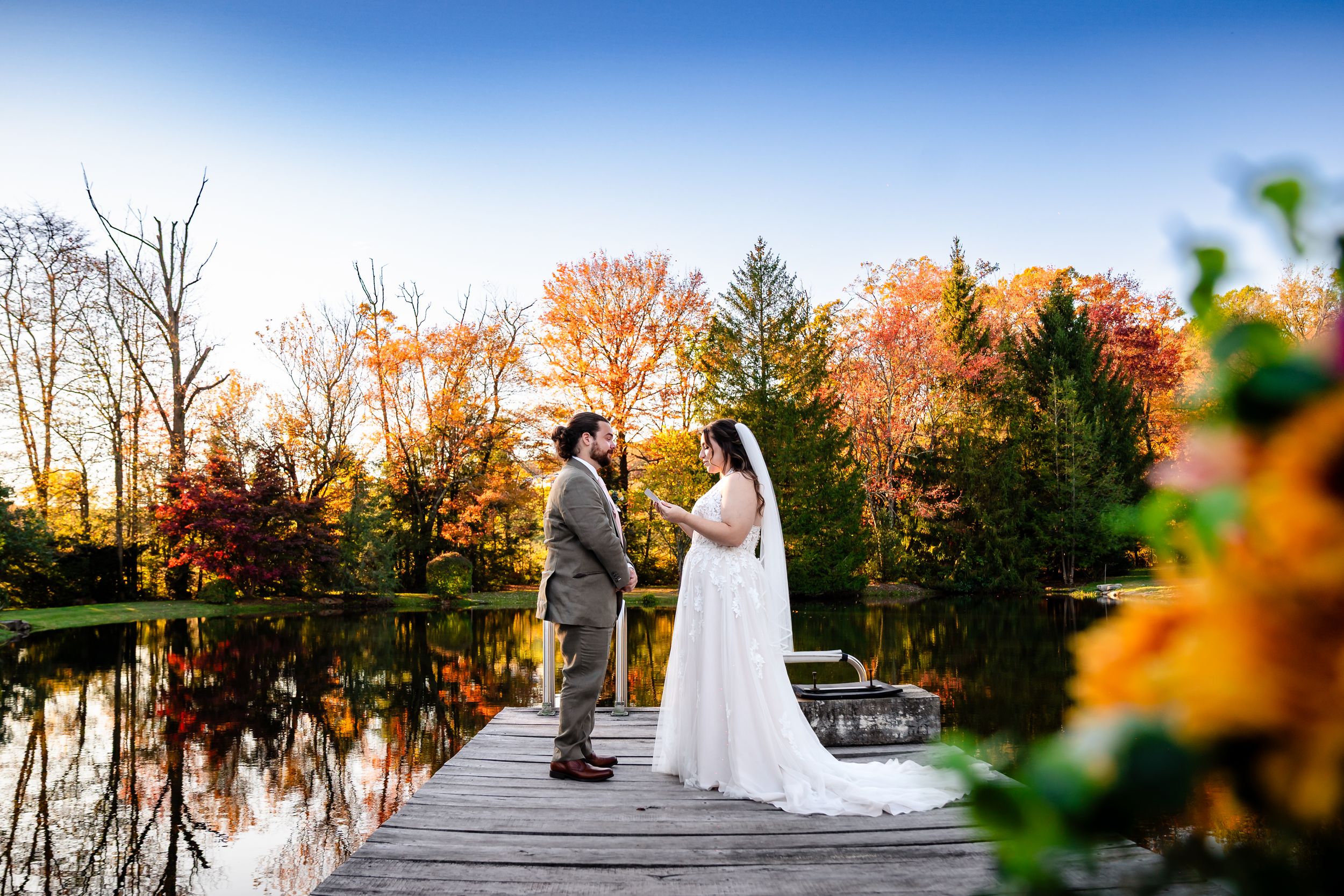 A wedding couple poses against vibrant autumn foliage and a tranquil lake during their fall wedding photoshoot.