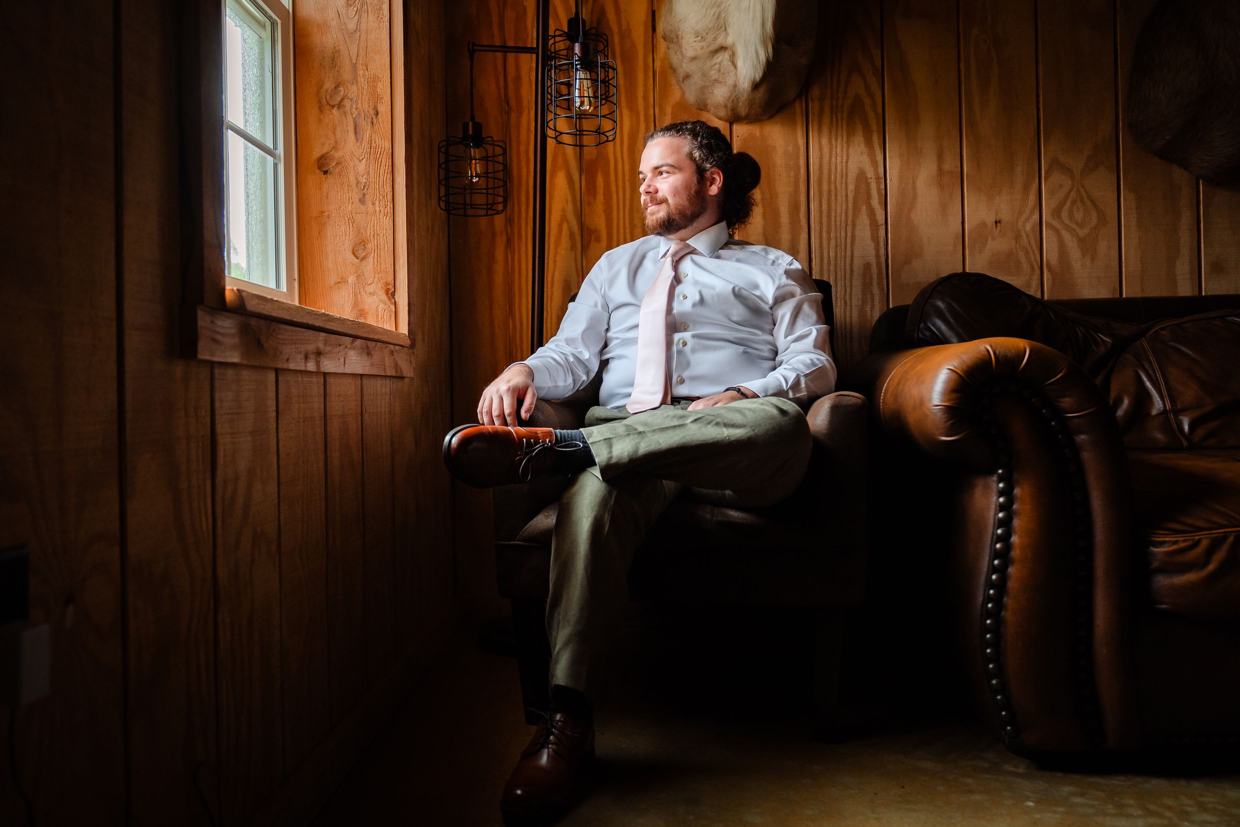 Person in business attire lounges casually on vintage leather couch in wood-paneled room with window light.