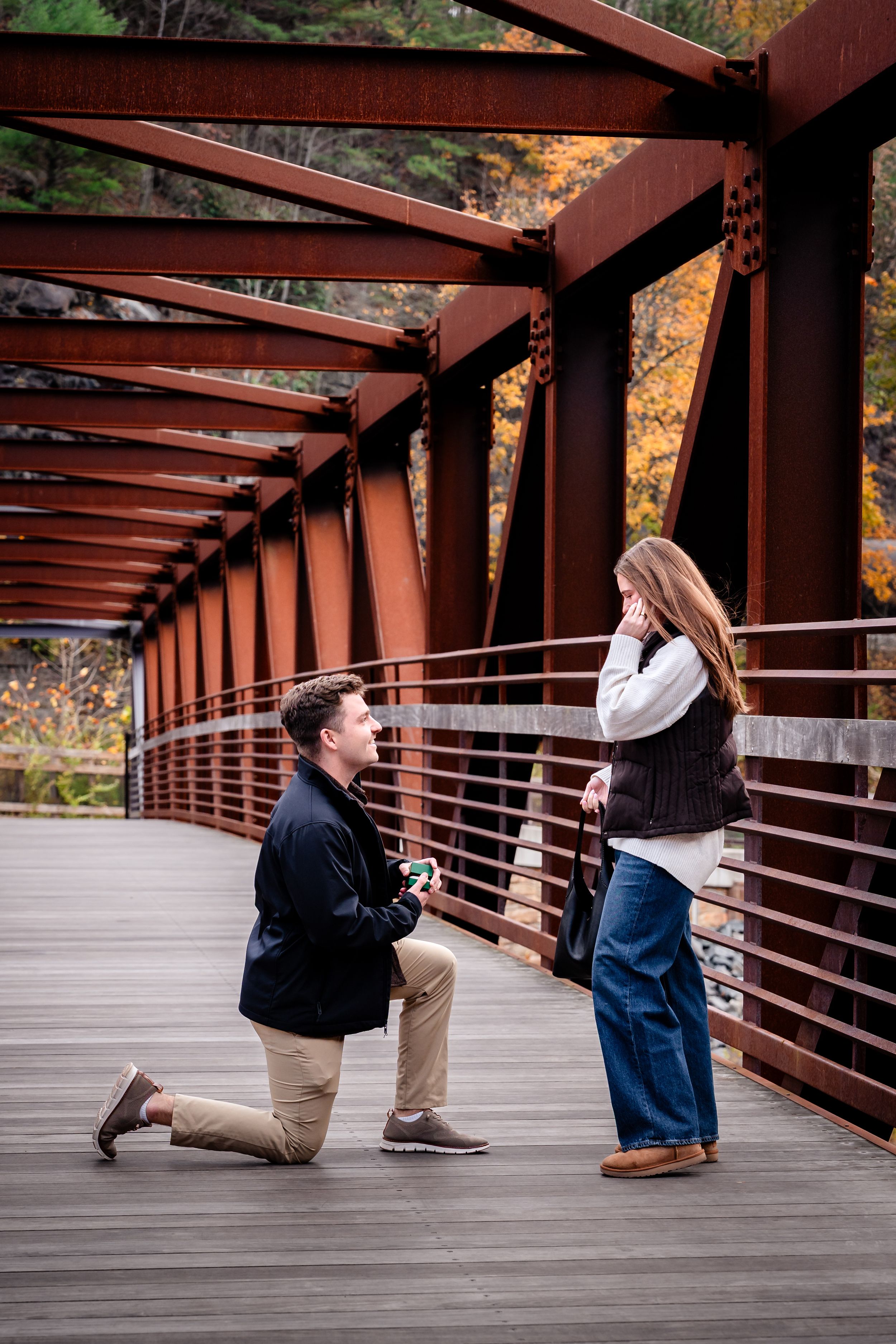 A romantic proposal moment on a rustic red bridge walkway surrounded by autumn foliage.